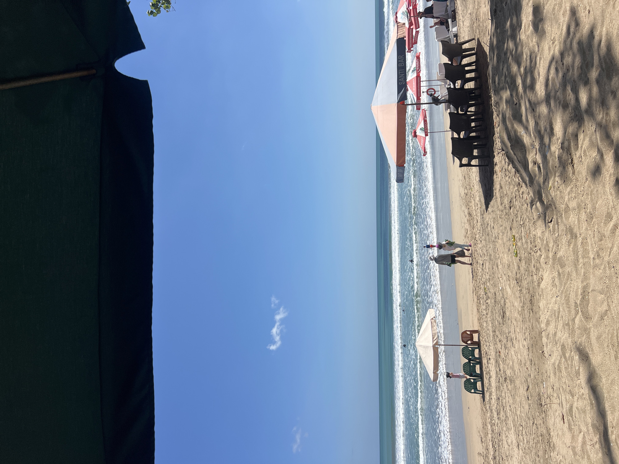 The image depicts a beach scene in Kuta, Indonesia, under a clear blue sky. In the foreground, a dark green umbrella partially obscures the top of the frame, casting a shadow on the sand. The beach itself is sandy, with tire tracks visible. Scattered on the sand are several plastic chairs, some arranged in rows, and a few beach umbrellas. One umbrella, with orange and gray panels, stands prominently, displaying the text "SANTI BAR." Two smaller red and white umbrellas with the text "LESSO" are also visible.
Further back, a line of waves breaks on the shore. Several surfers are in the water, identifiable by their dark wetsuits against the turquoise sea. On the wet sand near the water's edge, a few people are walking. Among them, a couple appears to be strolling, with one individual wearing a hat and carrying a bag. A child is also seen walking in the distance. The overall atmosphere is relaxed, suggesting a typical day at the beach.