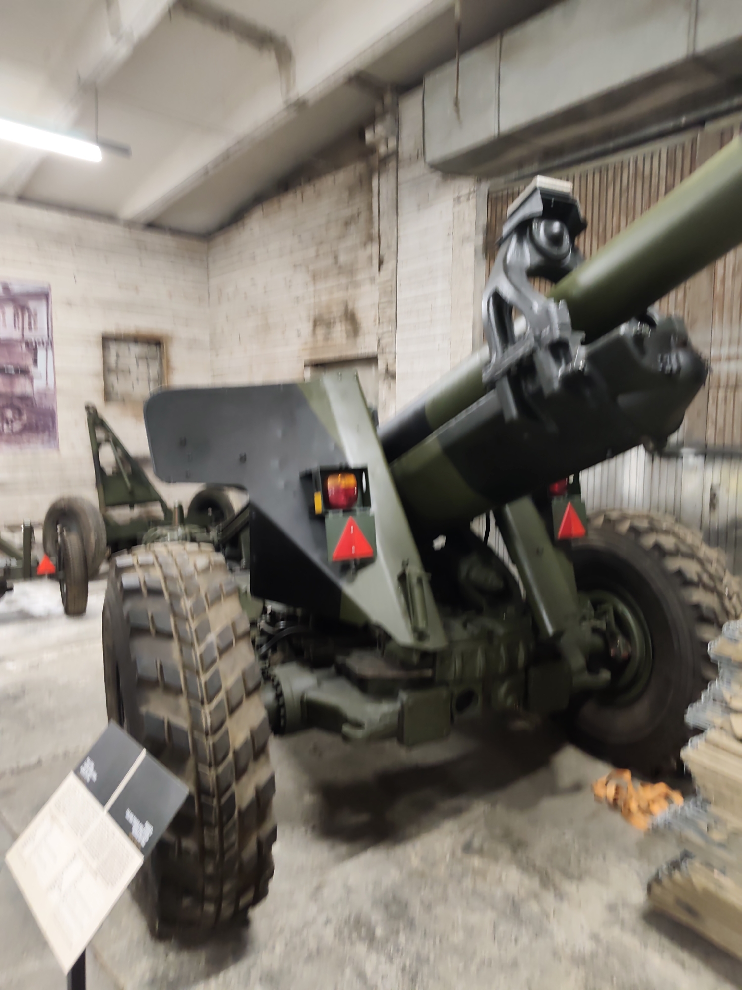 The image shows a close-up view of a large, wheeled artillery piece, likely a howitzer, displayed indoors. The weapon is painted in a camouflage pattern of dark green and black. The foreground is dominated by a massive, knobby tire, with the barrel and part of the carriage visible behind it. The barrel is a dull green and appears to be elevated. A metal support structure is visible beneath the barrel. To the left of the tire, a small informational sign on a black stand is partially visible. The background consists of a brick wall with some grimy patches, and wooden paneling further back. The ceiling has exposed beams and industrial lighting. The overall impression is of a museum or exhibition setting, likely dedicated to military history. The specific location in Viimsi, Estonia is not evident from the visual information alone.