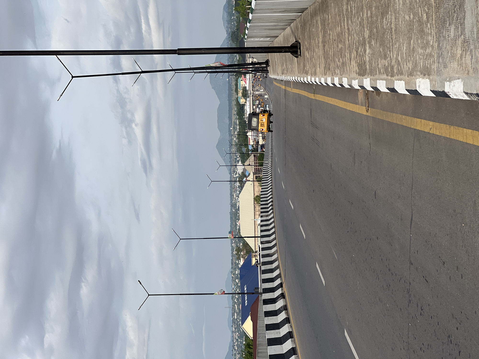 The image shows a highway overpass in Jalingo, Nigeria. The road is paved with asphalt and marked with white dashed lines. A yellow three-wheeled auto-rickshaw, known locally as a "Keke," is traveling away from the camera on the right lane of the road. Streetlights with a Y-shaped design are positioned along the sides of the overpass. The overpass is bordered by a concrete barrier with alternating black and white stripes. In the background, a city skyline is visible, with buildings and mountains under a cloudy sky. Two flags, one green, white, and red, and another with similar colors, are attached to poles along the overpass. The scene suggests a typical day in Jalingo, with traffic on the highway and the cityscape in the distance.