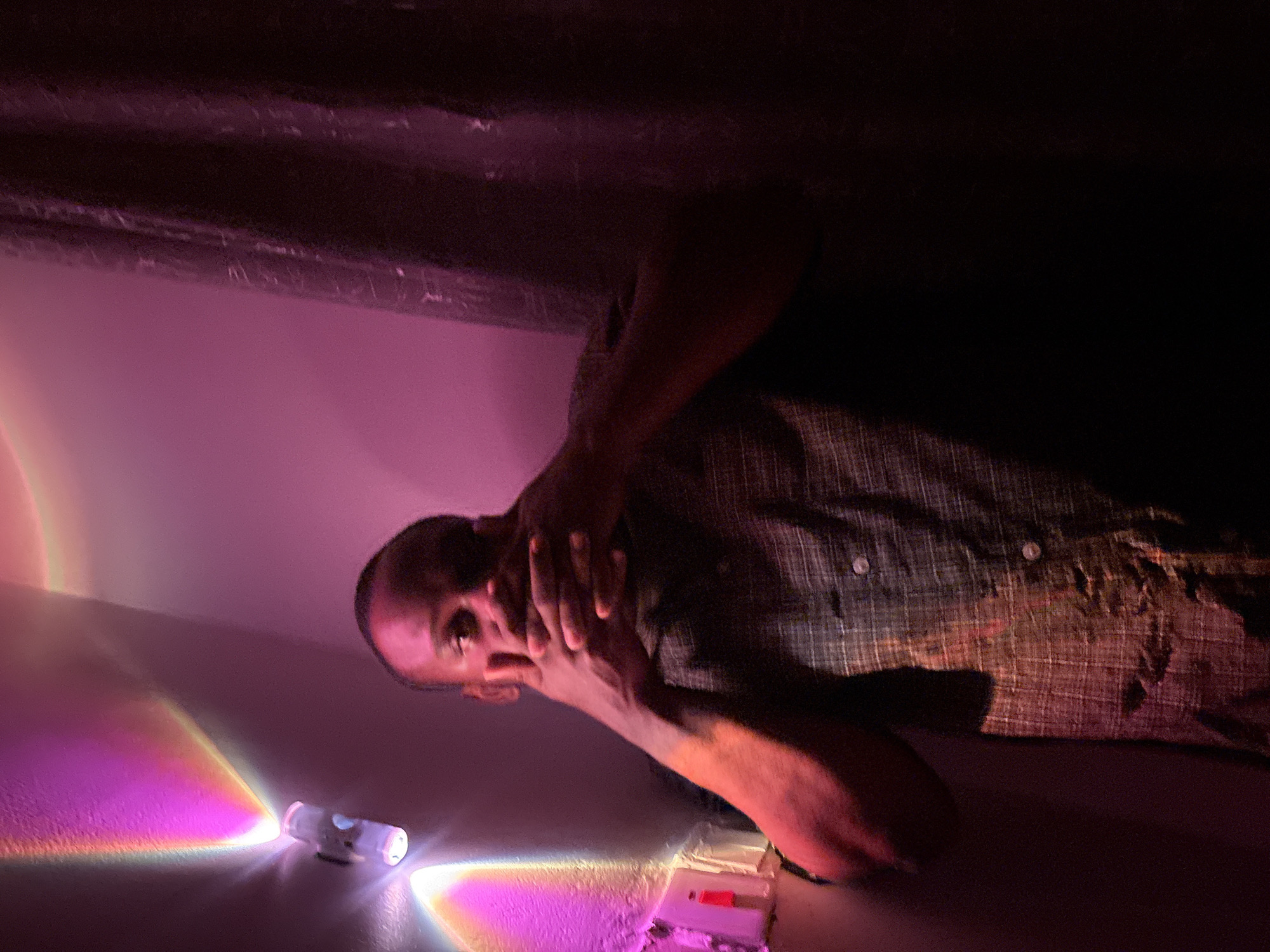 A person with dark skin, wearing a plaid shirt, is crouching in a low-lit space illuminated with pink and white light. The person's face is partially obscured by their hands. A small, cylindrical light source is visible on a surface reflecting a rainbow spectrum. Other small rectangular objects are also visible on the surface near the light. The location is unknown, and there is no indication this photo was taken in Iddo Tudunwada, Nigeria.