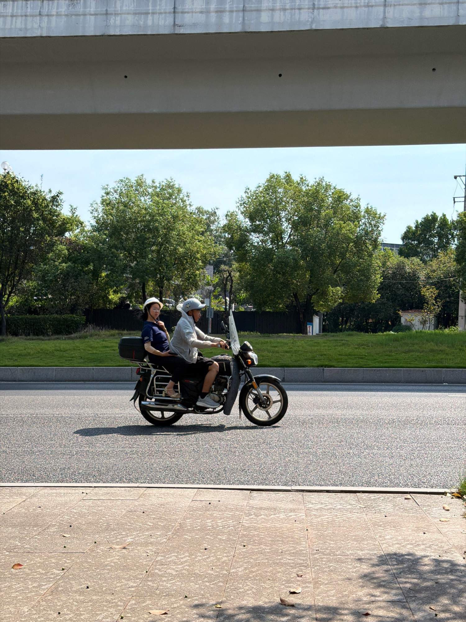 Two individuals are riding a motorcycle on a paved road in Jiangjin District, Chongqing, China. The rider, a man wearing a light-colored jacket and a helmet, is operating the motorcycle. The passenger, a woman in a dark blue shirt and a white helmet, is seated behind him. The motorcycle is black and has a windshield. The road is lined with a grassy median and trees. Above the scene, a concrete overpass casts a shadow. The sky is clear and blue. The image appears to have been taken during daylight hours.