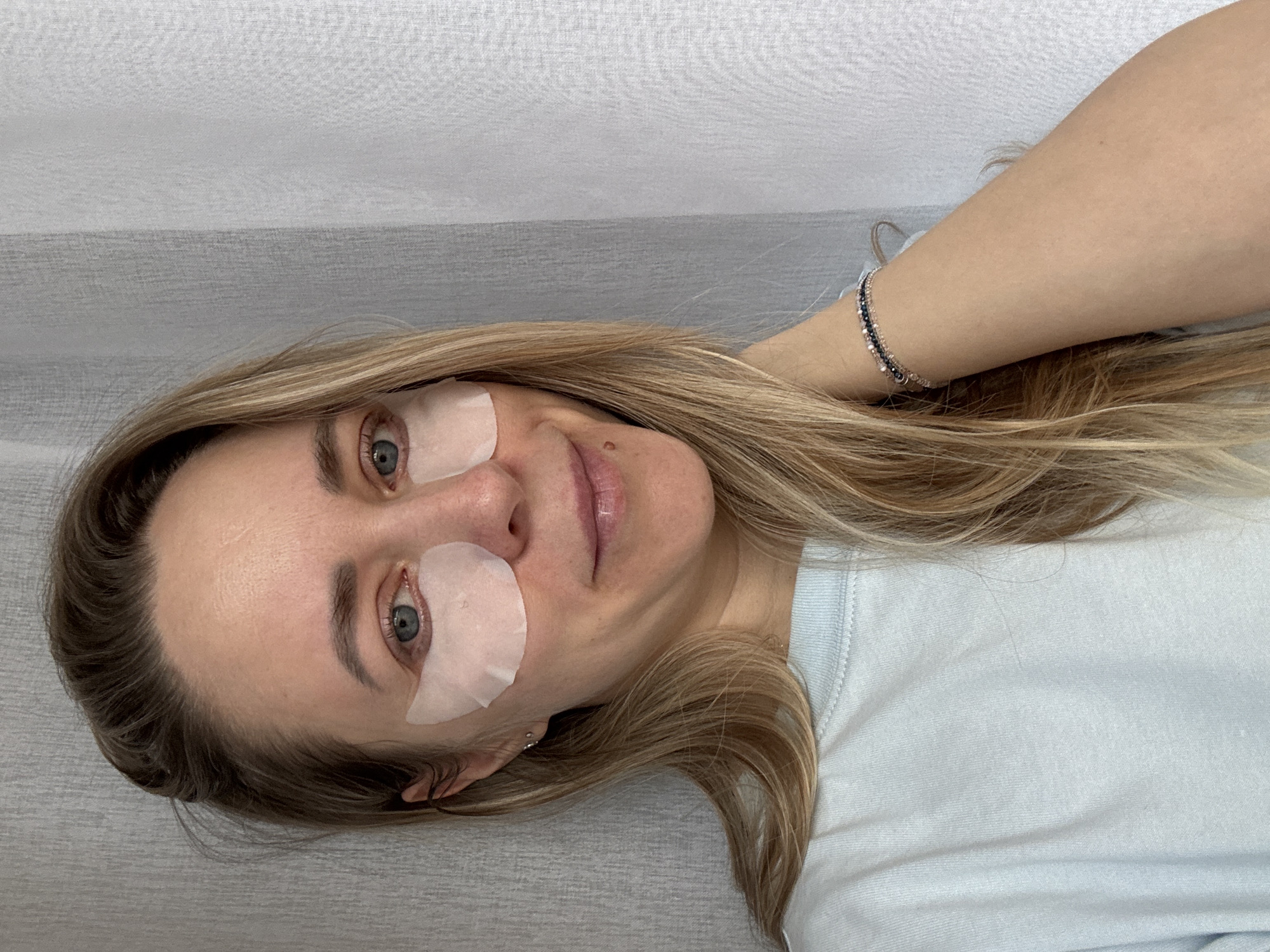 A woman with long blonde hair is lying down wearing eye patches under both eyes. She is wearing a light blue t-shirt and a silver bracelet on her wrist. The background is a light gray wall. The location is unspecified but presumed to be Chaikovsky, Russia based on a prior association. No further detail can be determined about subjects, objects, actions, interactions, or the environment.