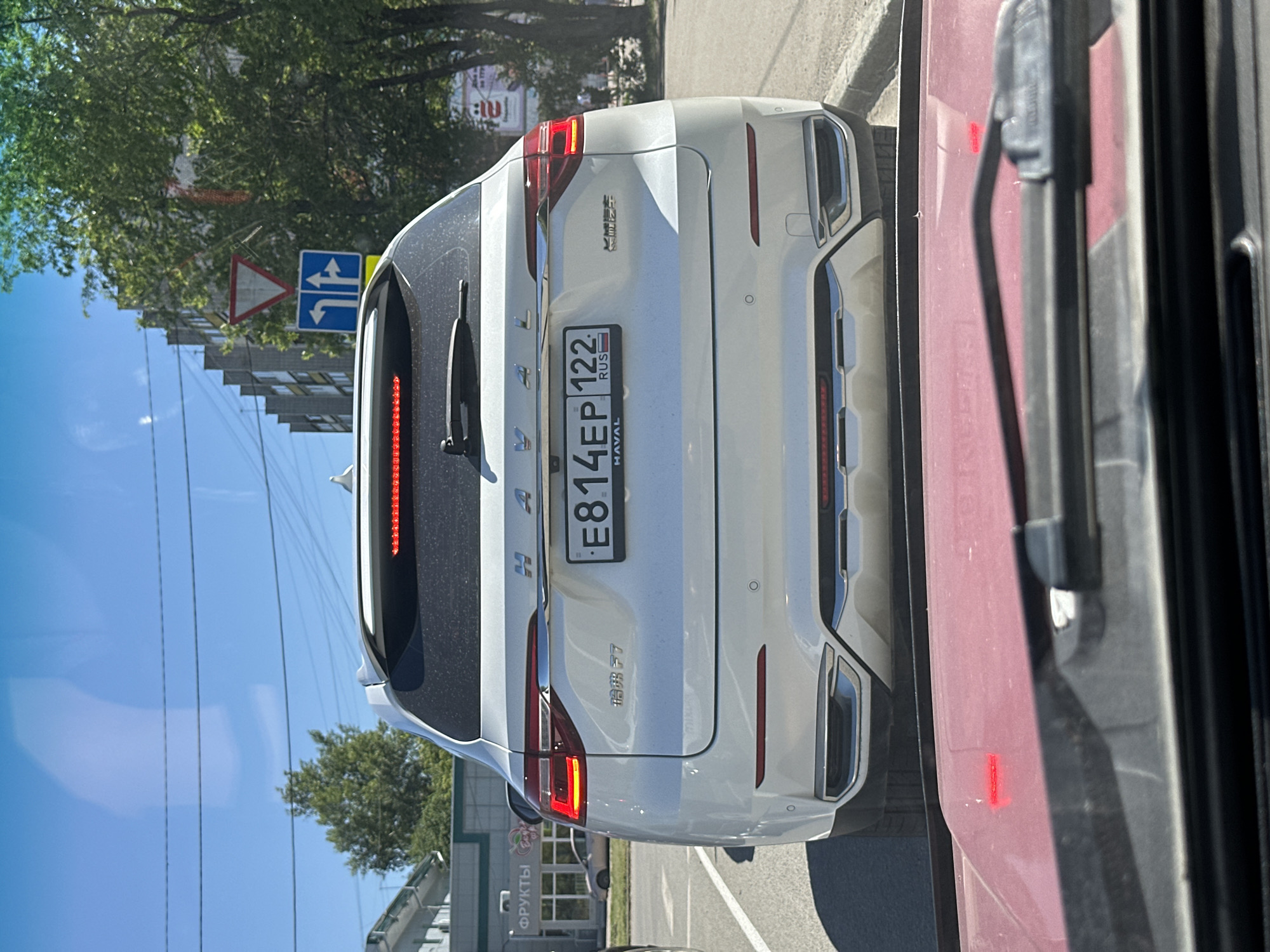 The image shows the rear view of a white Haval F7 SUV in Novoaltaysk, Russia. The car is parked on a paved road, with a red vehicle's windshield visible in the foreground, suggesting the photo was taken from inside another car. The Haval's license plate reads "E814EP 122 RUS". Behind the car, there are trees, buildings, and a blue traffic sign with arrows indicating directions and a yield triangle above it. The sky is clear and blue. A store with "ФРУКТЫ" (Fruits) signage is visible to the left of the Haval.