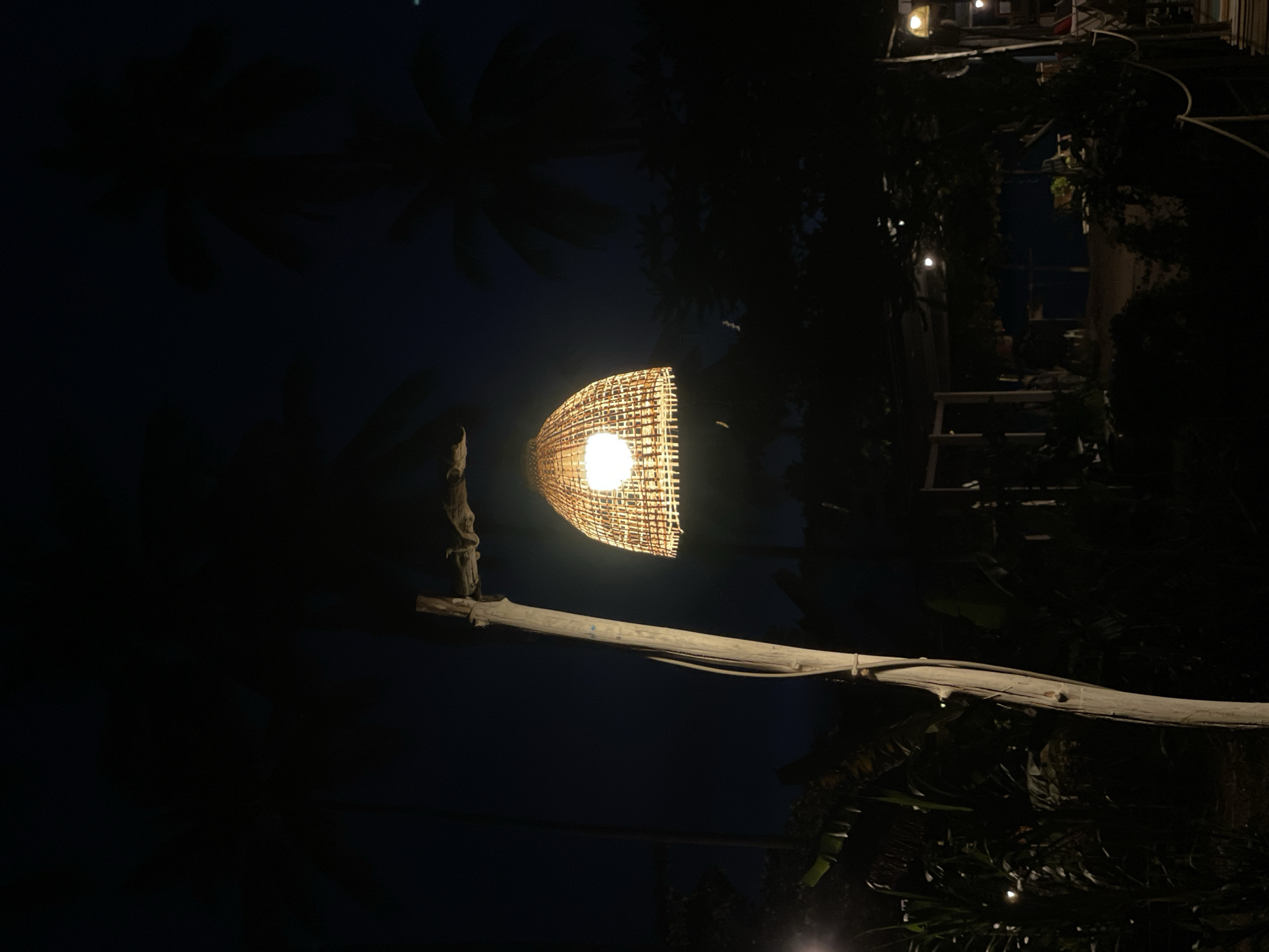 The image depicts an outdoor night scene, centered on a luminous light fixture. The fixture features a bowl-shaped lampshade meticulously crafted from woven natural material, possibly bamboo or rattan, which emits a bright, warm yellow light. This light source is suspended from a sturdy, rustic wooden pole or tree branch. A horizontal wooden beam extends from this main support, contributing to the rustic aesthetic.
The background is predominantly dark, with the distinct silhouettes of large tropical leaves or palm fronds visible against the deep blue-black night sky. In the distance, particularly towards the right side of the frame, several smaller, glowing lights can be observed, suggesting other structures or elements of a resort or outdoor area, partially obscured by more dark vegetation.
The time of day is clearly night, with artificial lighting being the primary source of illumination, creating a peaceful and ambient atmosphere. No individuals are present, and no specific activities are taking place. The overall setting, characterized by natural materials, tropical foliage, and evening lighting, strongly suggests a tranquil outdoor space, consistent with the provided location context of Baan Taling Ngam, Thailand. There is no visible text in the image.