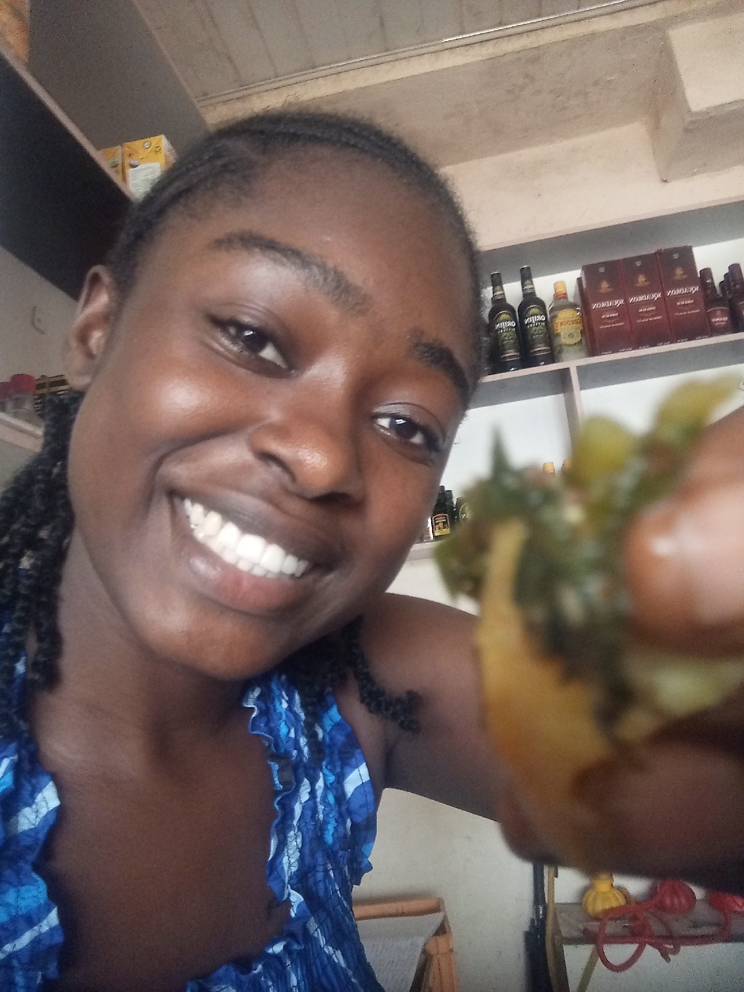 A close-up, slightly blurry selfie features a young Black woman with braided hair, smiling broadly and holding a piece of food up to the camera. She is wearing a blue and white patterned top. In the background, shelves are visible, stocked with various bottles, boxes, and other items, suggesting an indoor retail or storage environment. The lighting is dim and appears to come from overhead. The overall impression is casual and informal.