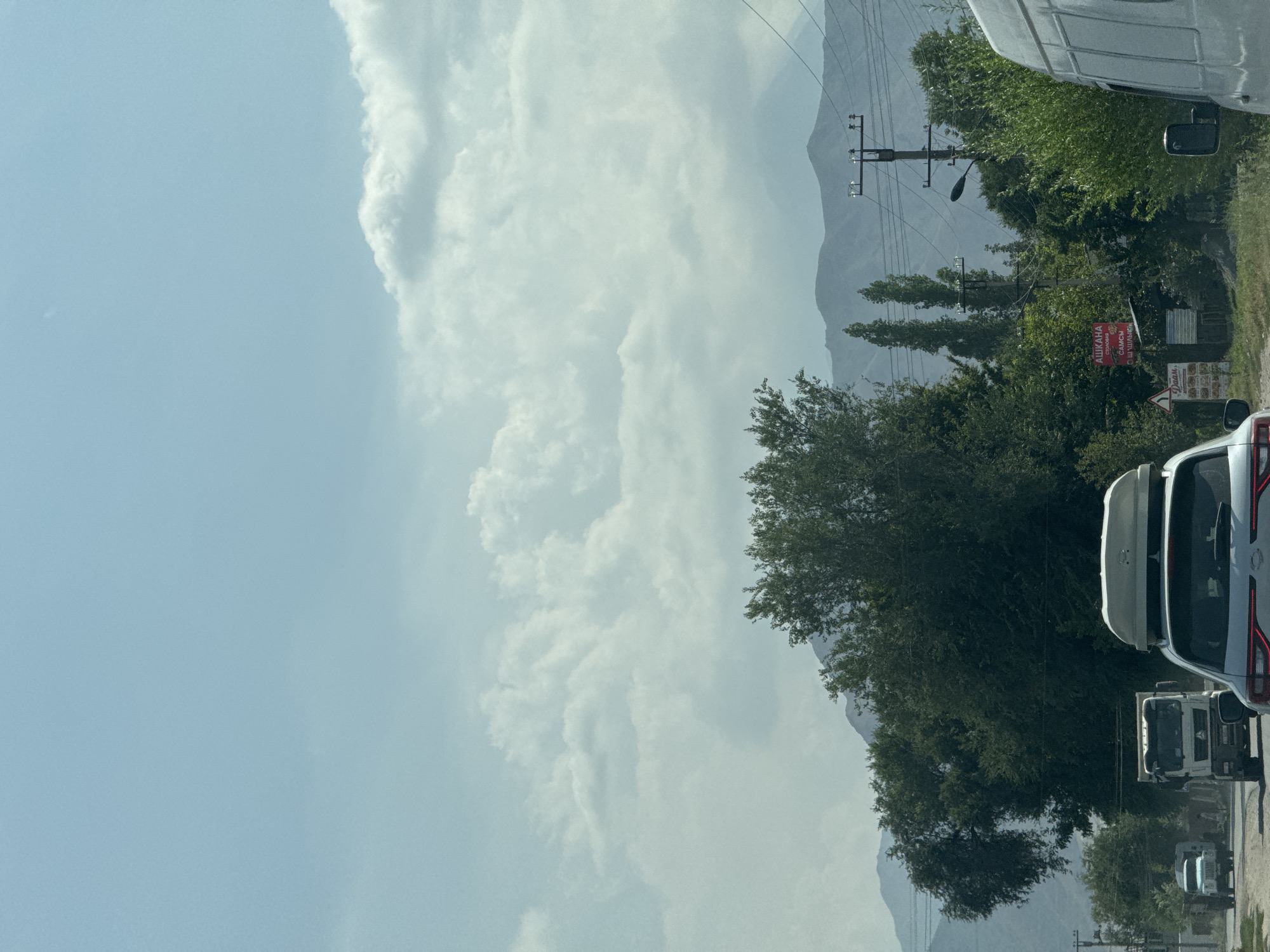 The image depicts a roadside scene in Beş-Küŋgöy, Kyrgyzstan, under a partly cloudy sky with distant mountains. In the foreground, a white SUV with a roof box is stopped in traffic. To the left, a white truck is also in traffic. Further back, a smaller white vehicle is visible. A red sign with Cyrillic text that appears to say "АШКАНА" (restaurant) is posted on a pole to the right of the road. Telephone poles with wires run along the side of the road, interspersed with dense green trees and vegetation. The overall impression is that of a rural or semi-urban setting in a mountainous region.