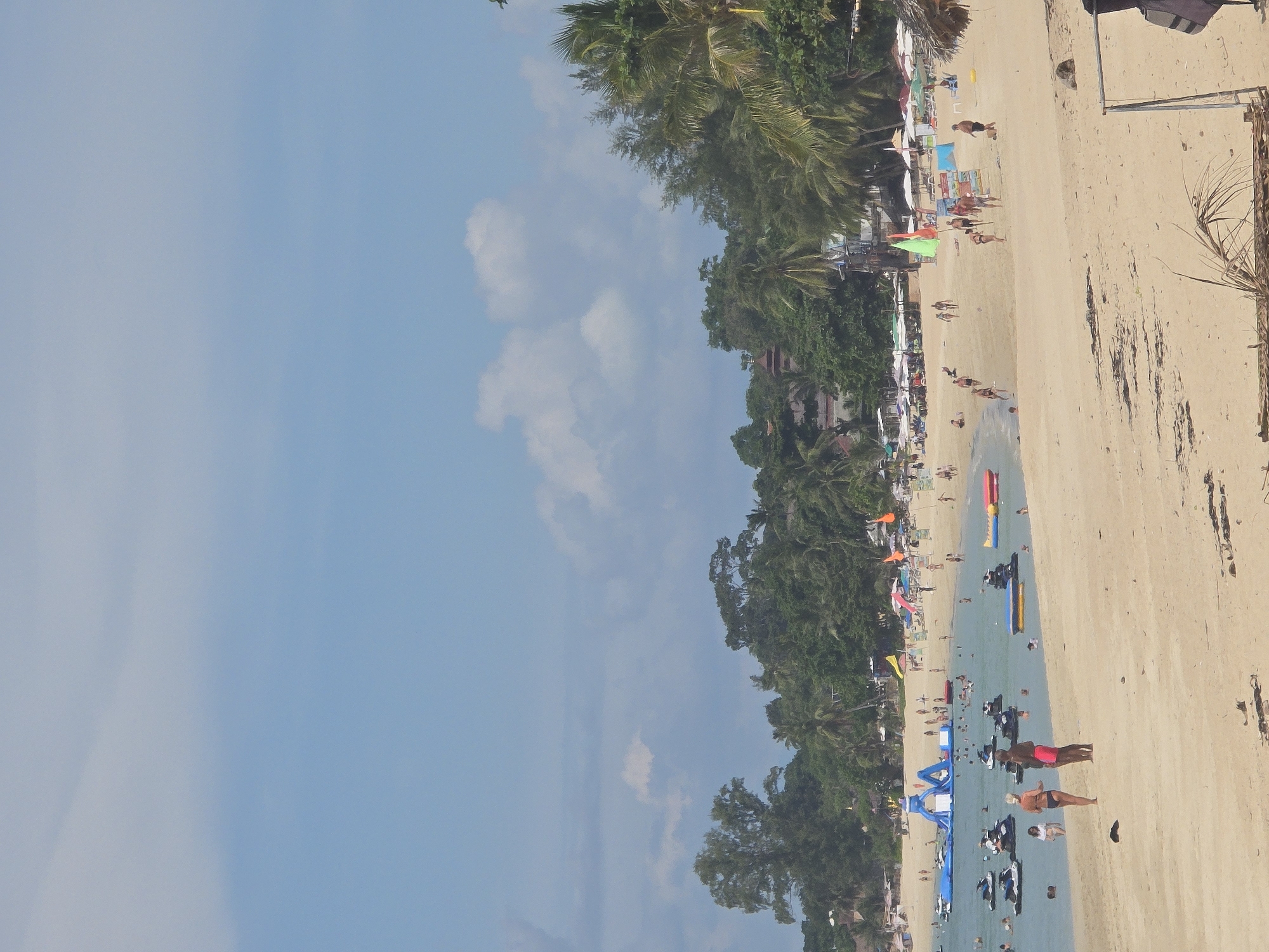 The image depicts a busy beach scene in Baan Lamai, Thailand. The foreground is dominated by a sandy beach with a few scattered pieces of dried foliage. In the mid-ground, a group of people are visible on the sand, with two individuals prominently standing in the center, one wearing a bikini and the other red shorts. Further into the scene, the ocean is populated with numerous jet skis and several people swimming or wading in the water. A large inflatable water park is set up in the sea, featuring blue slides and platforms. The background shows a lush green treeline with palm trees, typical of a tropical environment. Beach umbrellas and structures are scattered along the shoreline, indicating tourist amenities. The sky is a clear blue with a few scattered white clouds.
