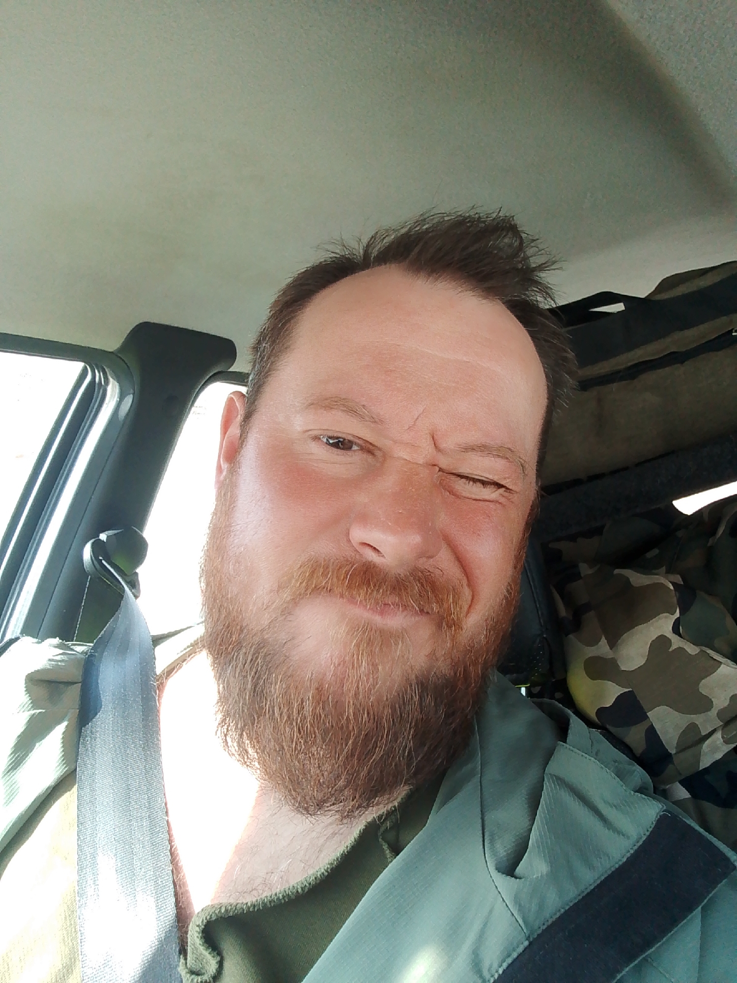 A man with a reddish beard and brown hair is in the driver's seat of a vehicle. He is winking at the camera. He is wearing a green jacket and a seatbelt. There is a camouflage bag or backpack behind him. The scene is indoors, likely inside the vehicle. The location is not determined by the image.