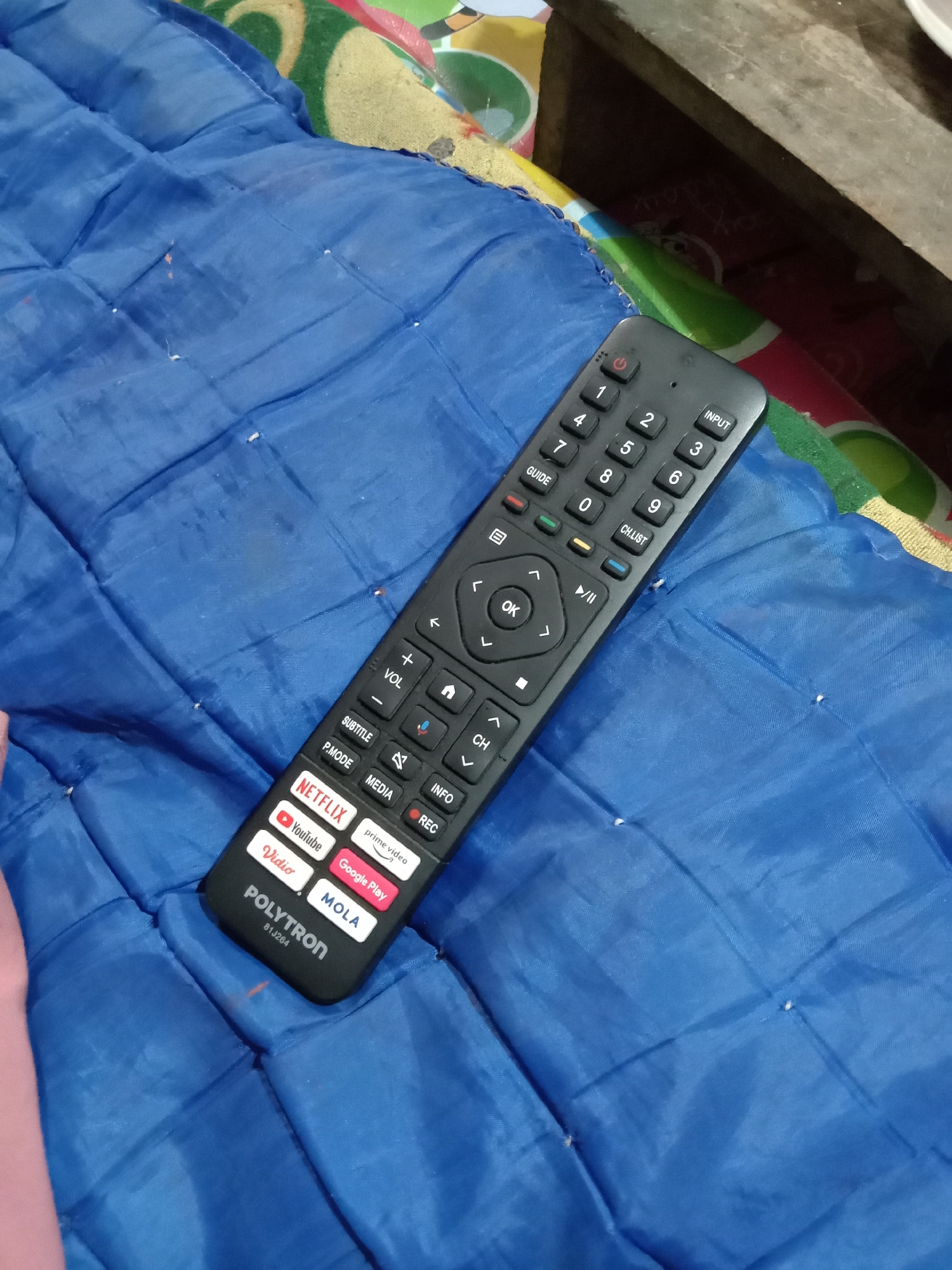 A black television remote control rests on a blue quilted fabric surface. The remote features various buttons including numbers 0-9, directional arrows with an OK button in the center, playback controls, and shortcut buttons for streaming services like Netflix, YouTube, Prime Video, Vidio, MOLA, and Google Play. The brand "POLYTRON" and a model number "81J2G4" are visible on the lower part of the remote. The blue fabric is textured and appears to be part of a blanket or mat. The background is out of focus but suggests a rustic indoor setting with hints of other patterned fabrics and wooden surfaces. There are no people present in the image. The image is taken from a slightly elevated angle, looking down at the remote. The overall scene is indoors, likely a living space.