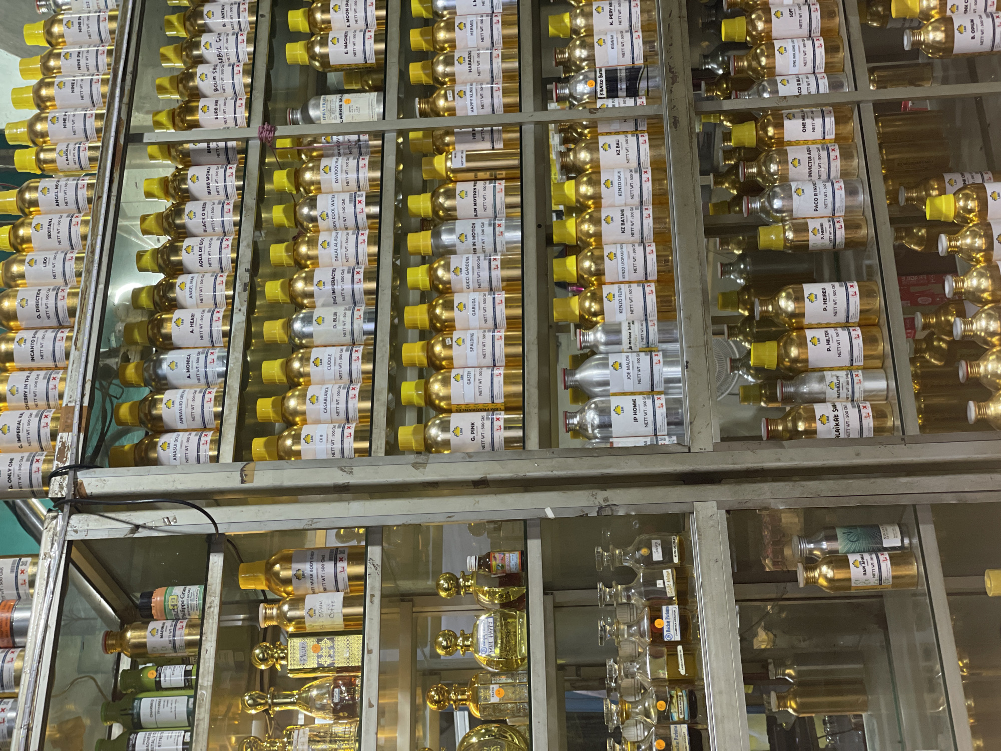 The image displays a wall of shelves filled with numerous bottles, likely perfumes or essential oils, arranged in a retail setting. The bottles are primarily made of glass and many have a metallic gold or silver finish, with some clear glass bottles also present. They are neatly organized in rows and columns. Each bottle appears to have a label with text, suggesting they are branded products. Some labels are clearly visible, including brand names like "DIOR", "KENZO", and "CHANEL", along with product names and indications of net weight. The scene is brightly lit, likely due to overhead lighting in a shop or store. The shelves themselves are made of metal, with some showing signs of wear. The overall impression is of a well-stocked perfume or fragrance store. There are no people visible in the frame. The context suggests this is a commercial environment where fragrances are sold. The lighting suggests it is daytime or a well-lit indoor space.