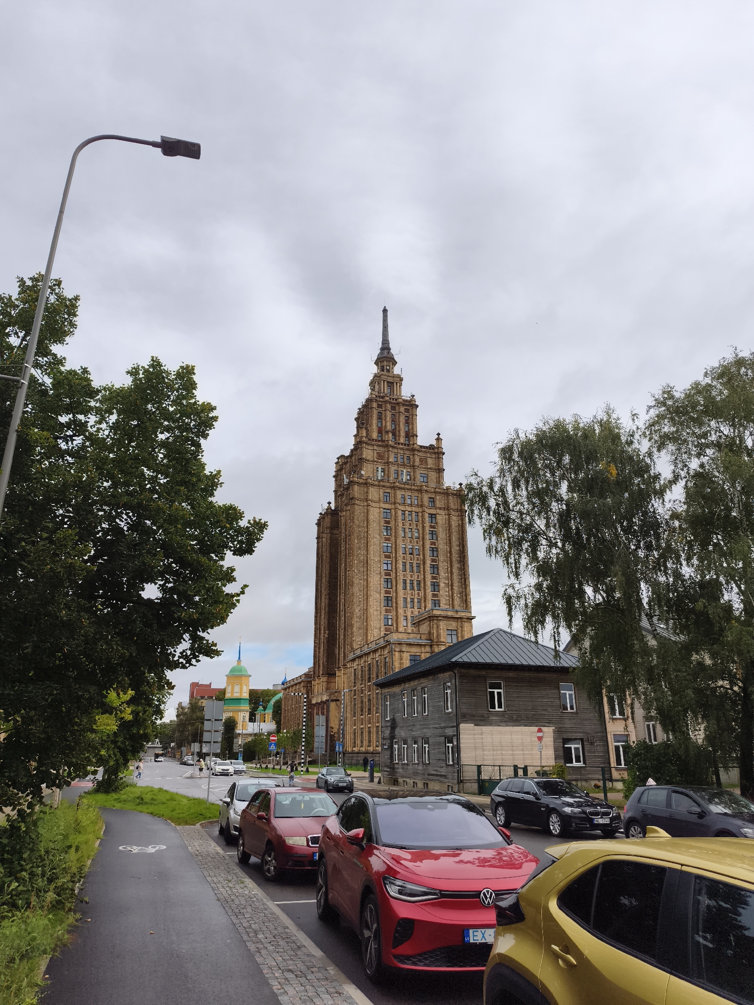 The image shows a street scene in Riga, Latvia, featuring the Academy of Sciences building, a prominent Stalinist Empire-style skyscraper constructed between 1953 and 1956. In the foreground, cars are parked along the street, including a red Volkswagen ID.4 and a yellow car. A bicycle lane is visible on the left side of the road. Trees line the street, and the sky is overcast. Other buildings, including a wooden house and a building with a green and yellow spire, are also visible. A lamppost stands on the left side of the image. The overall atmosphere is that of a city street on a cloudy day.