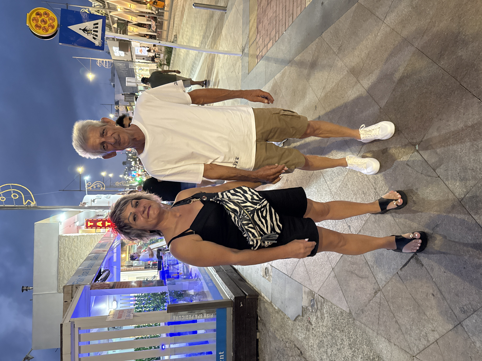 A couple, a man and a woman, stand on a paved sidewalk in Ayia Napa, Cyprus. The woman, on the left, has short, greyish-brown hair and wears a black slip dress and black sandals. She carries a black and white zebra-print crossbody bag. The man, on the right, has short, grey hair and wears a white t-shirt and beige cargo shorts. He has on white sneakers. Behind them, shops line the street, illuminated by string lights and the setting sun. A pedestrian crossing sign is visible to the man's right. The sky is a deep blue, indicating dusk.