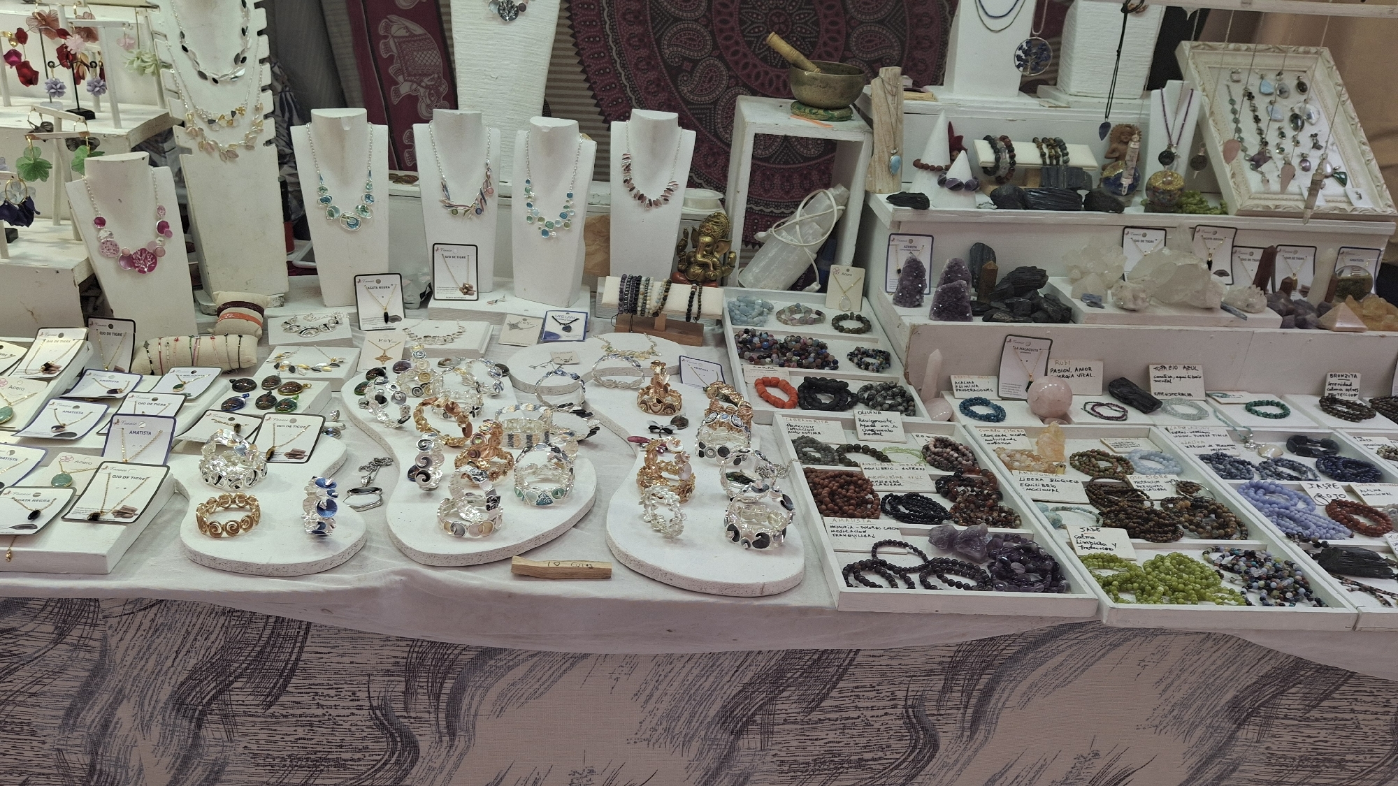 The image shows a display of jewelry and crystals at an outdoor market, likely in La Fresneda / la Freixneda, Spain. The stall is set up on a table covered with a patterned cloth. The display includes various necklaces, bracelets, and rings, as well as raw crystals and polished stones. The jewelry is presented on white display stands and in trays. The items vary in material, with some appearing to be made of metal, beads, and semi-precious stones. Several cards with handwritten labels are visible, likely indicating prices or types of stones. In the background, a tapestry with a pattern is visible, along with a small brass bowl and a statue of a deity. The overall impression is a vendor at a market showcasing artisanal goods.