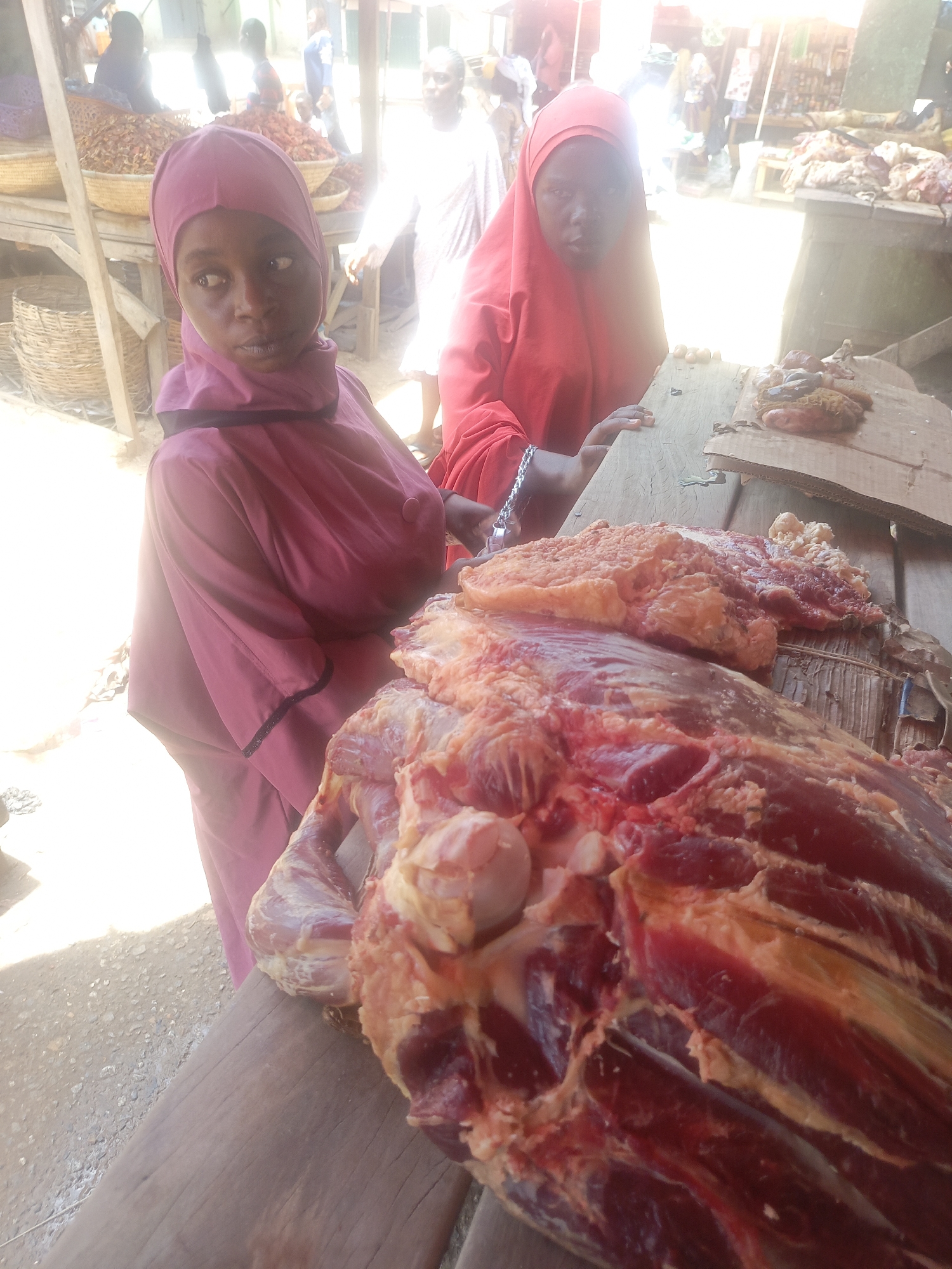 Two women wearing hijabs stand behind a wooden table laden with raw meat in what appears to be an outdoor market in Akwanga, Nigeria. The woman in the foreground is wearing a pink hijab and looks towards the left. The other woman, wearing a red hijab, stands slightly behind and to the right. The table is covered with a large cut of meat. There are other market stalls and people in the blurred background.