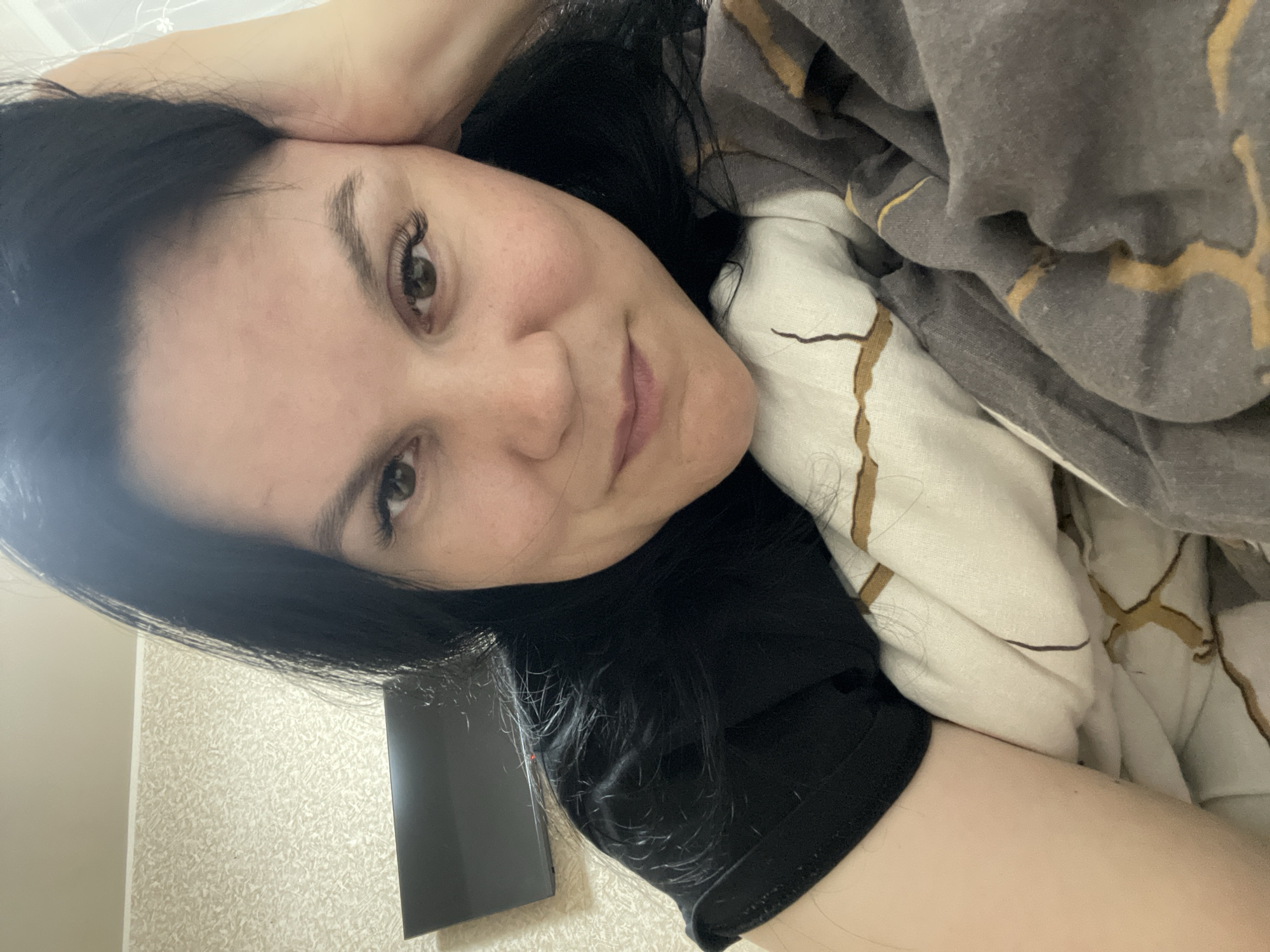 A woman with dark hair and light skin is lying in a bed. She has brown eyes and is looking at the camera with a slight smile. Her head is resting on her hand. She is covered in a bed sheet, which has a brown and white pattern. A dark smartphone is near her black t-shirt. The photo was taken indoors. The scene's location in the city of Izhevsk, Russia, is unknown.