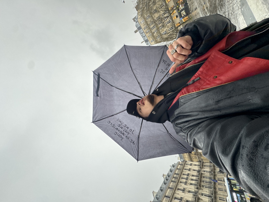 An individual, appearing male, is depicted in a low-angle outdoor portrait holding an open dark grey umbrella against an overcast, light grey sky. The subject wears a black baseball cap, a black hooded sweatshirt, and a black leather jacket, with a red garment visible underneath the jacket on their right side. Multiple rings adorn their left hand, including a prominent ring with a large stone on the ring finger. The individual's head is tilted upwards, with the mouth slightly open, and gaze directed towards the sky or umbrella. The umbrella's interior canopy displays inverted, white text. The scene is set in an urban environment in Paris, France, identifiable by the distinctive light-colored Haussmannian architecture of multi-story buildings with balconies visible in the background on both sides. The street appears wet, and a blue city bus is partially visible on the left side of the frame, indicating rain or recent precipitation.
