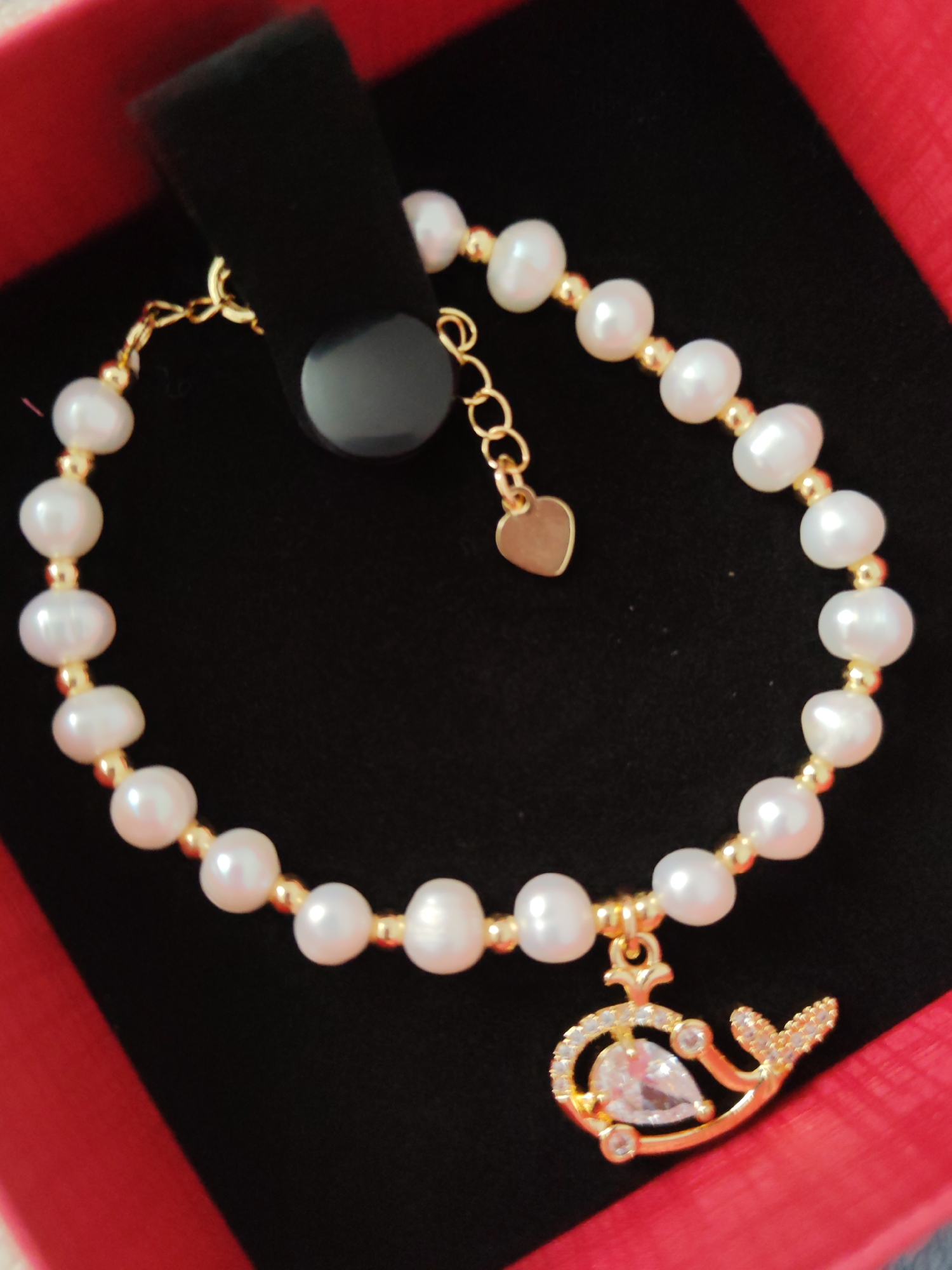 A close-up, overhead shot features a pearl and gold bracelet with a whale charm, resting on black velvet within a red jewelry box. The bracelet consists of numerous irregularly shaped white pearls interspersed with small, round gold beads. A delicate gold chain extends from one end of the bracelet, adorned with a small, flat heart-shaped charm. The focal point is a gold whale charm, intricately detailed with tiny clear stones, a larger pear-shaped clear stone at its center, and a textured tail. The bracelet is displayed against a black velvet background, which is itself inside a red, open jewelry box. The image is sharp and well-lit, highlighting the textures and details of the jewelry. The location of Komsomolsk-on-Amur, Russia is not discernible from the image.