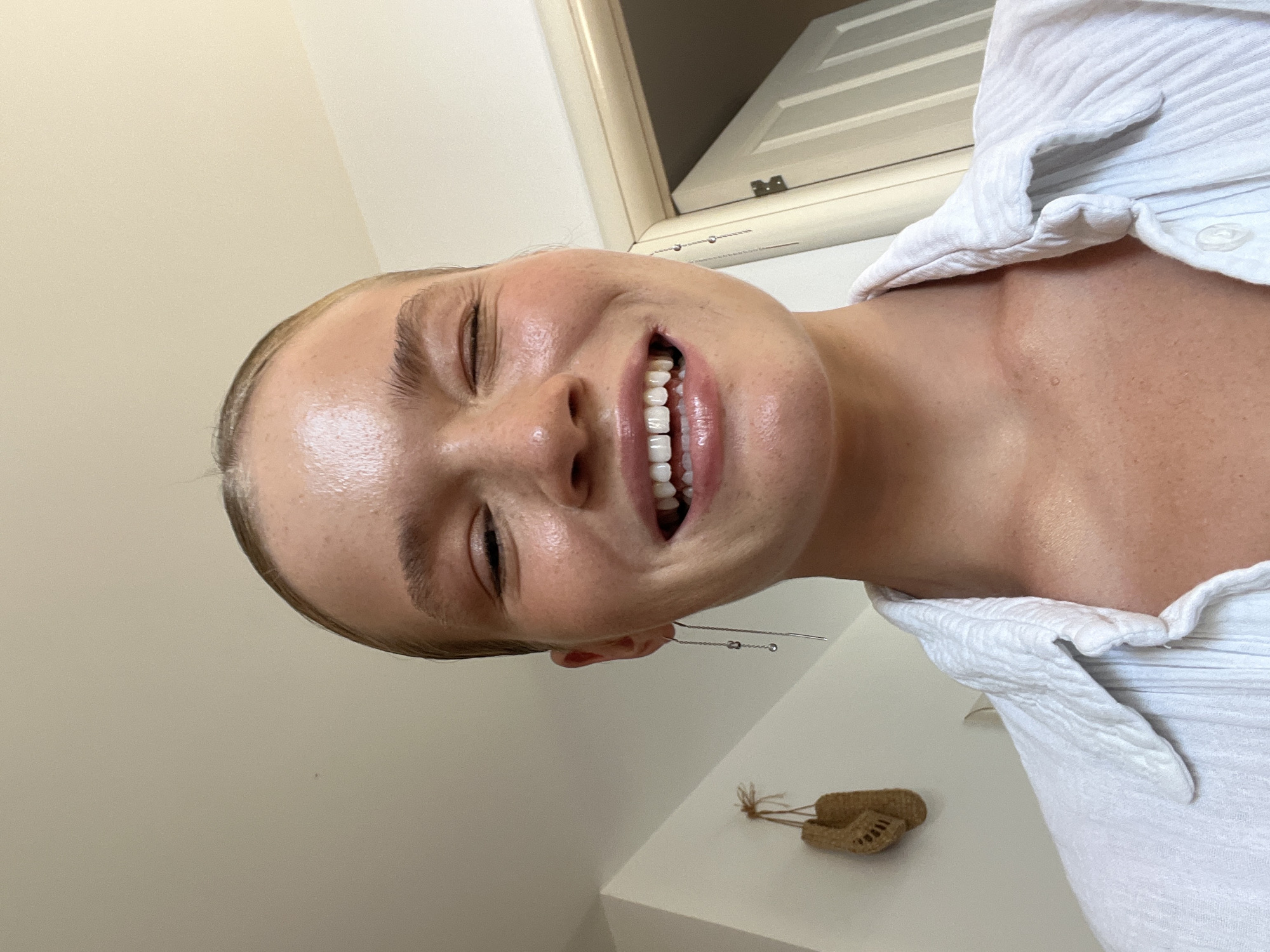 A young woman with fair skin and blonde hair tied back smiles with her eyes closed. She is wearing a white collared shirt. A long, delicate earring with small silver charms dangles from her visible ear. The background is a neutral wall with a door and a small woven decoration hanging on the wall. The lighting suggests it is daytime, with sunlight possibly coming from a window. The scene is likely indoors, possibly in a residential setting, and there is no indication of the city of Budva, Montenegro.