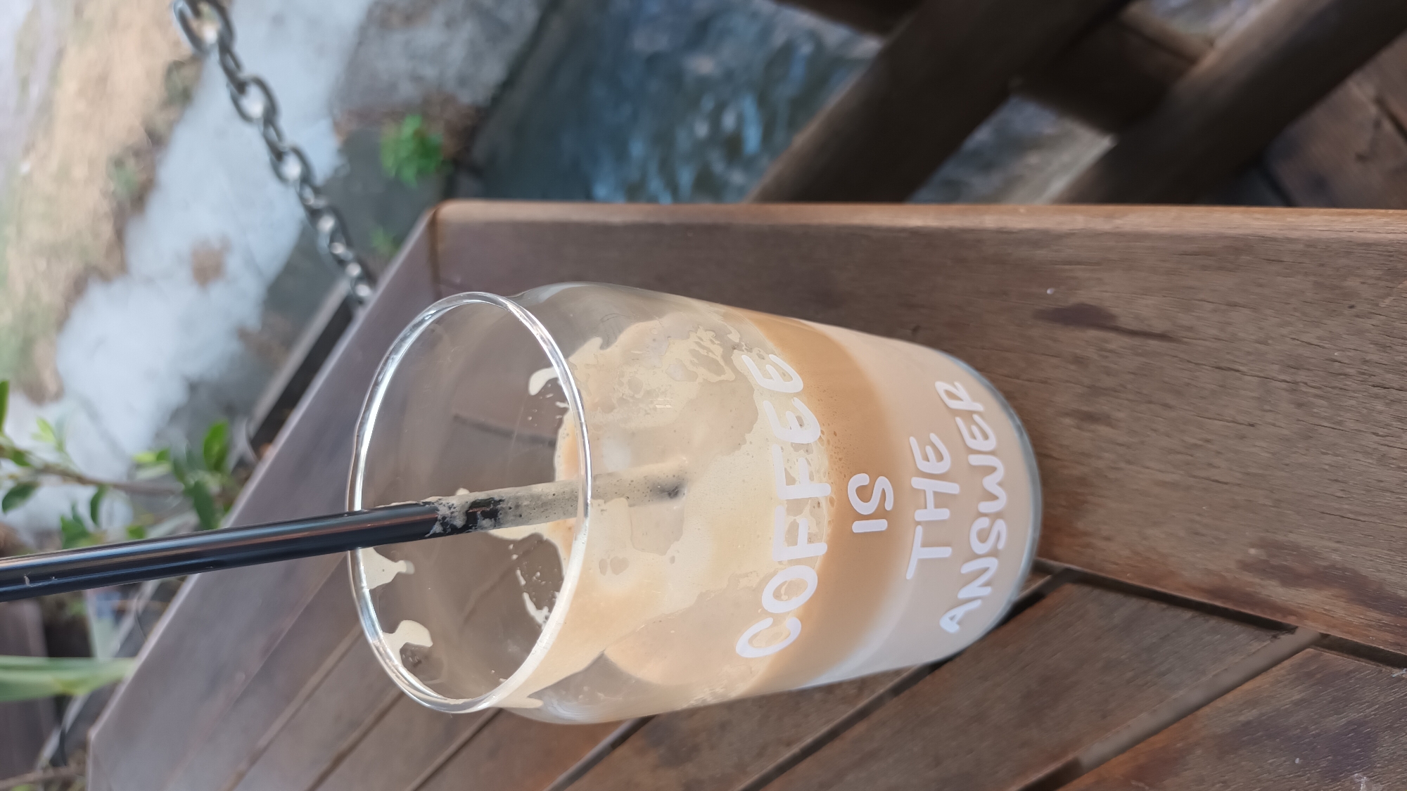 A glass with coffee and a straw sits on a wooden table. The glass has the words "COFFEE IS THE ANSWER" printed on it in white text. In the background, there is a chain link fence and a body of water. The image is taken from a slightly high angle, looking down at the glass. The lighting is natural, suggesting it is daytime. This scene appears to be outdoors, possibly at a cafe or restaurant with a view of a canal or river. The environment suggests a relaxed, possibly vacation setting in Vetren Dol, Bulgaria.