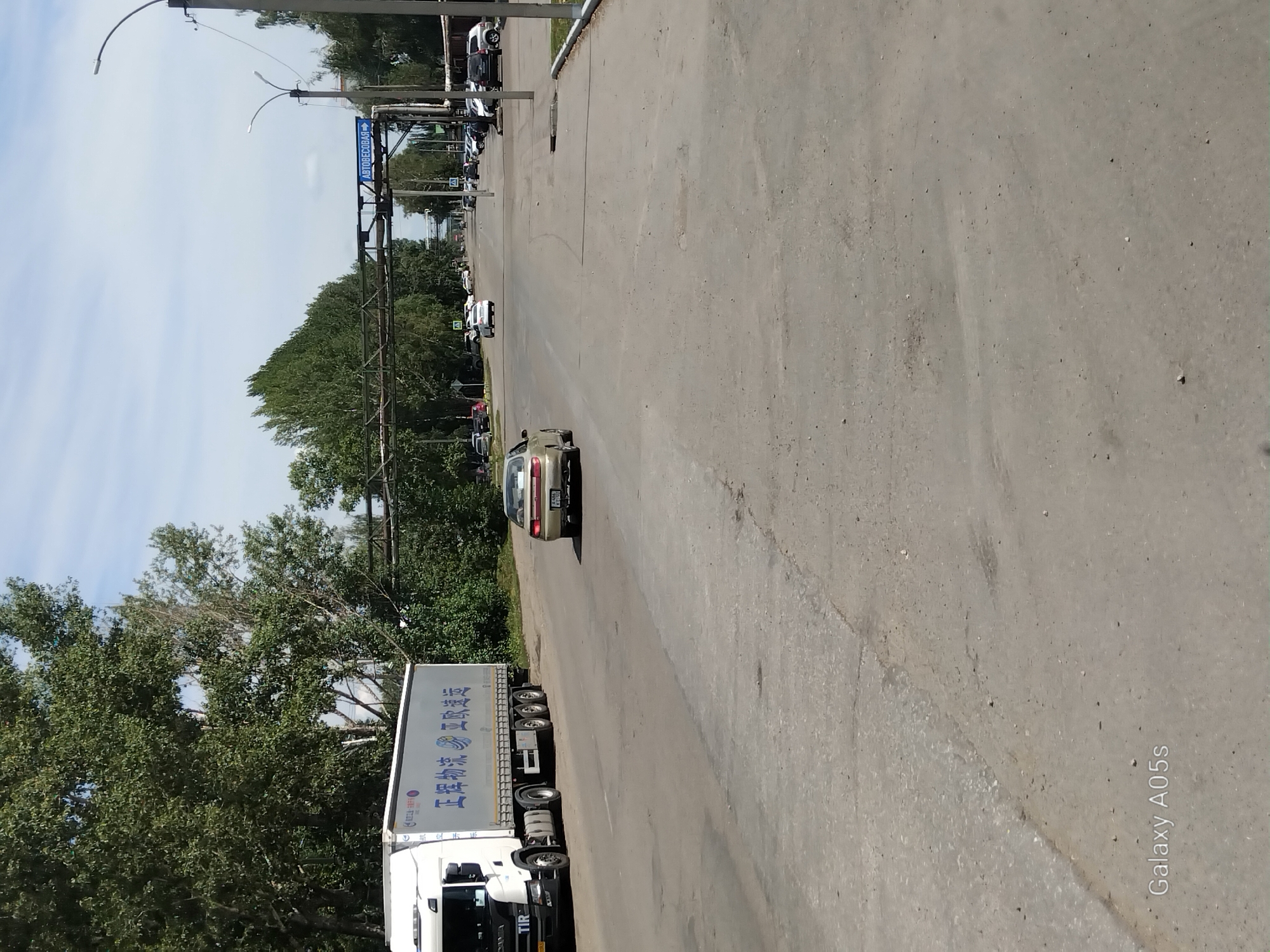 The image shows a street scene in Aleysk, Russia, with a white semi-trailer truck on the left side of the road. The truck has Chinese characters on its side. In the middle of the road, a beige sedan is driving away from the camera. The road is paved and appears to be in moderate condition with some patches. Trees line both sides of the road, and a blue sign with white lettering, "АВТОВЕСОВАЯ" (Autoweighing Station), is visible overhead on a metal structure. Several other vehicles are parked along the right side of the road. The sky is partly cloudy. The watermark "Galaxy A05s" is present in the bottom left corner.