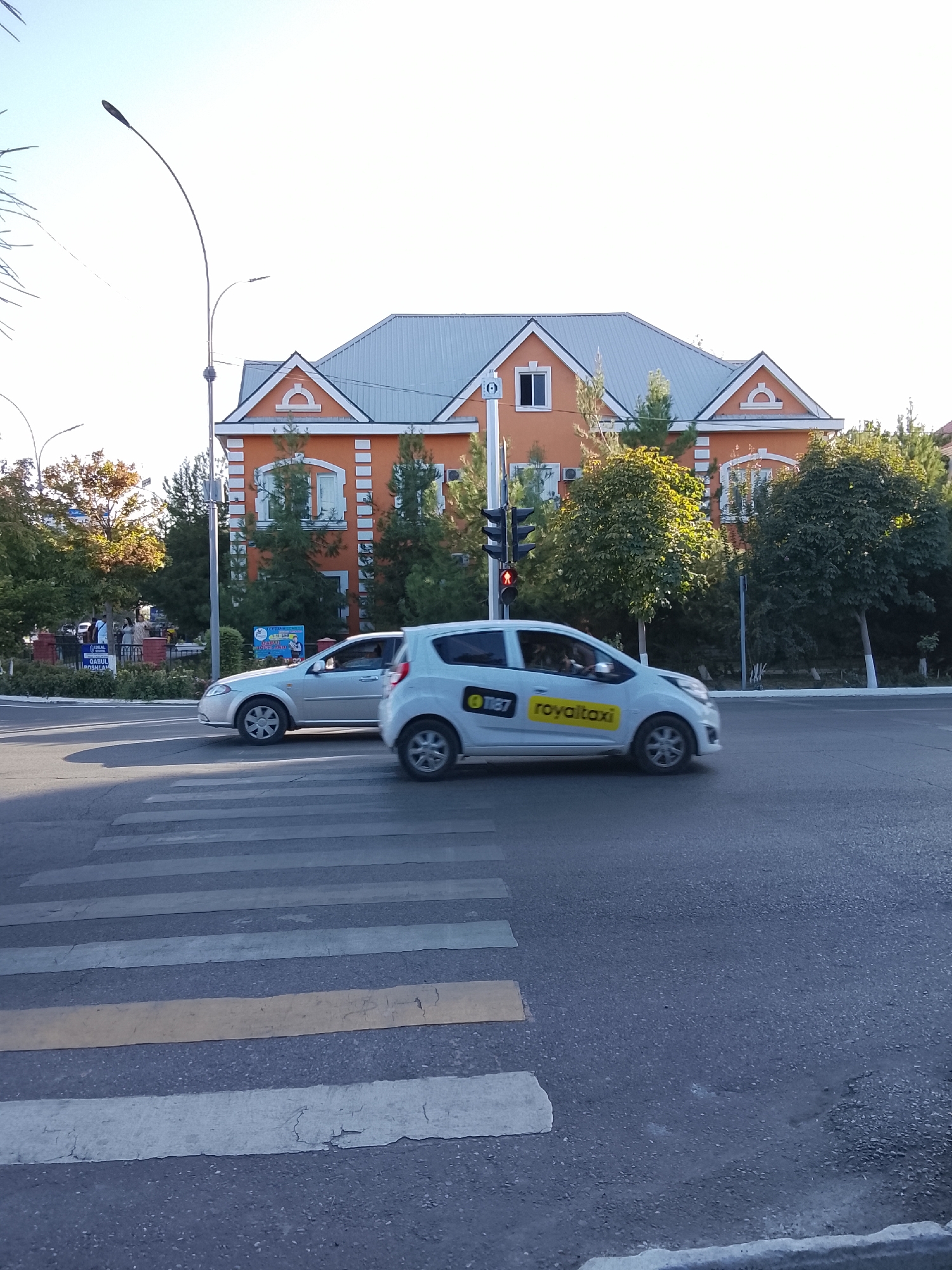 This is a daytime, eye-level, medium shot photograph taken on a street in Fergana City, Uzbekistan. In the foreground, a pedestrian crossing is visible on an asphalt road. Two cars are crossing the pedestrian crossing. The car on the left is a silver sedan, and the car on the right is a white hatchback with "royaltaxi" branding. Behind the cars, a bright orange building with a grey roof stands out. Several trees are planted in front of the building and along the street. A traffic light is visible on the left side of the road, showing a red light. The sky is clear and bright blue. The overall scene depicts a typical urban streetscape in Fergana City.