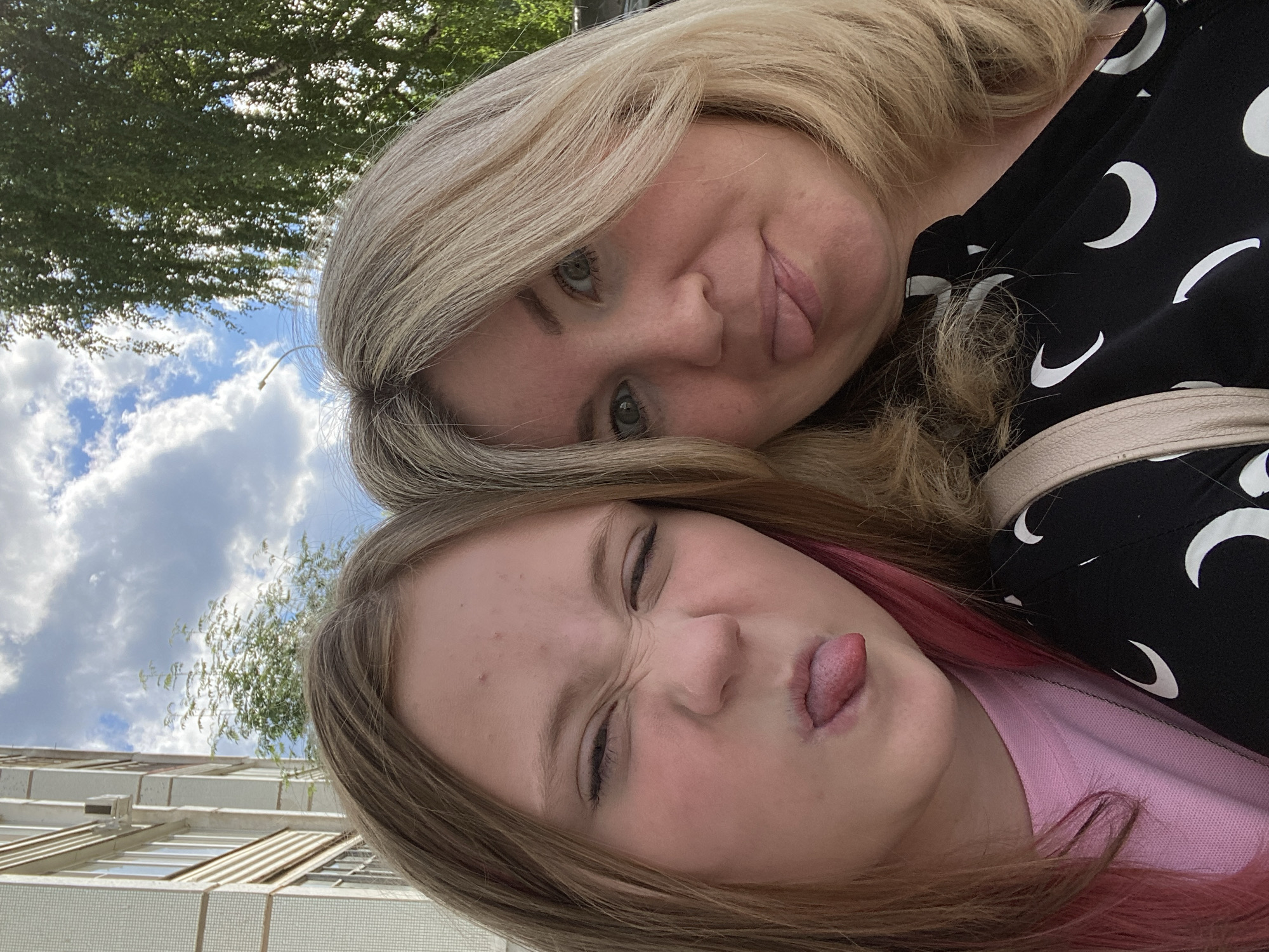 Two individuals, one younger with blonde hair streaked with pink and the other older with blonde hair, are taking a selfie outdoors. Both are sticking out their tongues and making playful faces. The younger person is wearing a pink shirt, while the older person is wearing a black shirt with a crescent moon pattern. Behind them, a building with a tiled facade is visible, along with trees and a cloudy blue sky. The scene appears to be in the city of Chaikovsky, Russia, based on the architectural style of the building which is typical of Soviet-era apartment blocks found in Russian cities.
