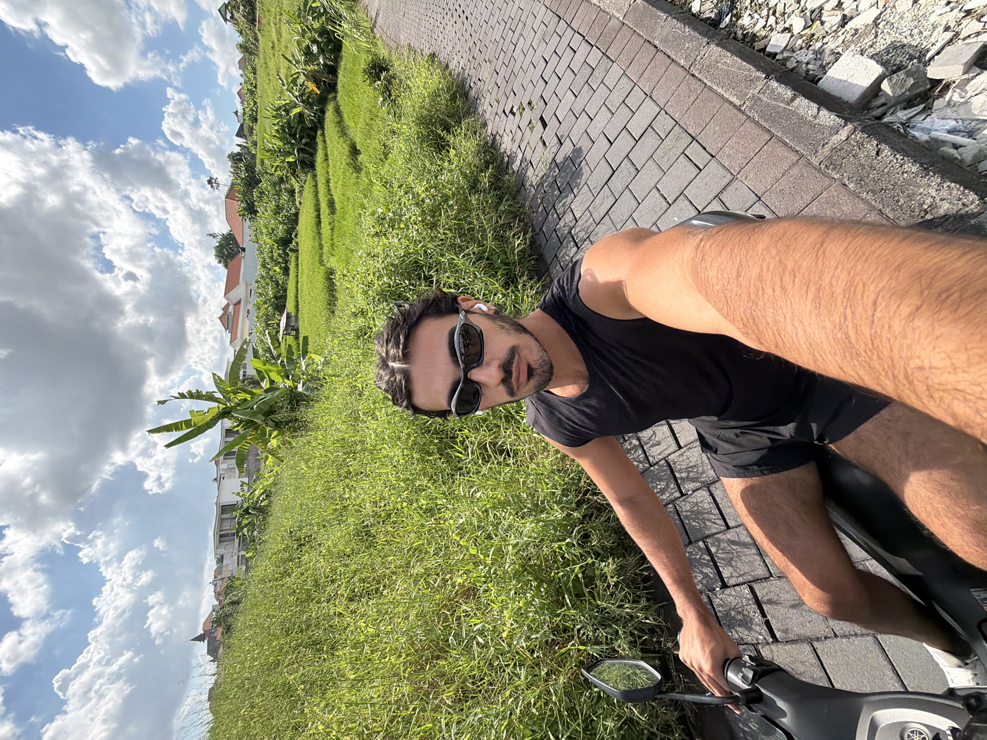A man wearing a black T-shirt and black shorts is sitting on a scooter on a brick path in what appears to be a lush, tropical area. He is wearing sunglasses and appears to be taking a selfie. The location is likely North Kuta, Indonesia, based on the presence of tropical vegetation and building styles in the background. To the left of the path is tall green grass, and in the background are houses and banana trees. The sky is partly cloudy.