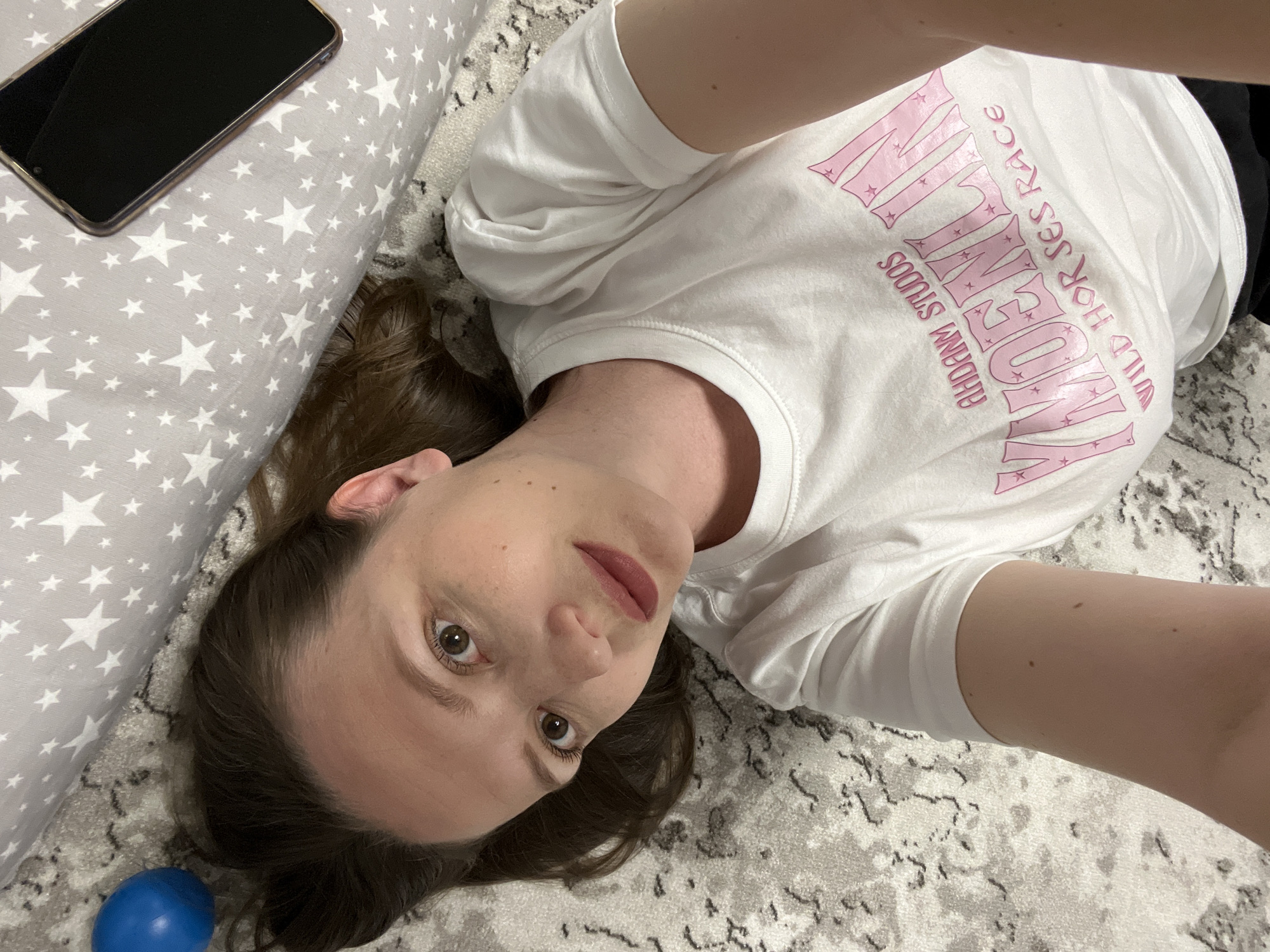 A woman is lying on a patterned rug, wearing a white t-shirt with pink lettering, in an indoor setting. A grey pillow with white stars and a black smartphone are adjacent to her. A blue ball is near her head. The location cannot be determined to be Chaikovsky, Russia based on the image.