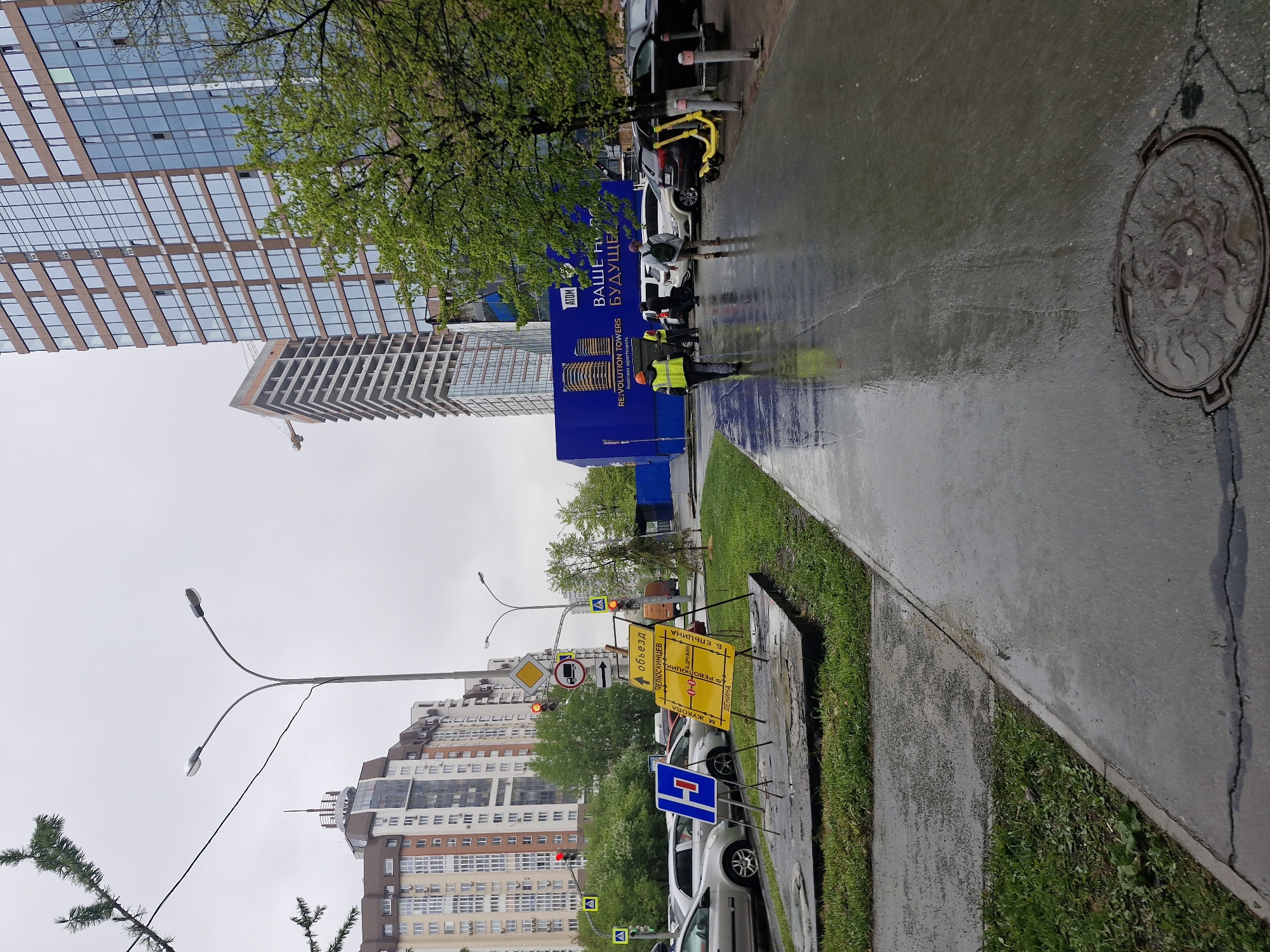 The photograph captures a city street scene in Yekaterinburg, Russia, under overcast weather conditions, likely after a rainfall, evident from the wet pavement. In the middle ground, two workers wearing bright yellow vests are positioned near a construction site hoarding. The hoarding, blue, promotes the "RE:VOLUTION TOWERS" business apartments.
On the right, the street is lined with a patch of green grass and a sidewalk. A manhole cover is visible in the upper right corner of the frame.
Further down the street, there are various road signs, including detour signs and a no-entry sign. Cars are parked along the roadside.
In the background, there are several buildings, including a tall modern skyscraper and apartment blocks. A street lamp is seen reaching upwards.
The overall impression is of an urban area undergoing construction activities.