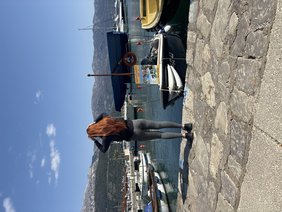 The image depicts a marina in Budva, Montenegro, under a clear blue sky with scattered white clouds. A single individual, seen from the back, stands on a paved quay composed of irregular grey flagstones. The person has long, wavy red hair, wears a black long-sleeved top, grey leggings, and black athletic shoes, with both hands raised to the head, seemingly adjusting their hair.
To the immediate right of the individual, a dark-hulled motorboat is docked in the calm, blue-green water. This boat features a blue canopy, a yellow-orange lifebuoy, and a vertical pole displaying a rectangular sign advertising "TRANSFER & PANORAMA BOAT" with images of coastal scenes and a Montenegrin phone number (062 68 763 890). The boat's registration number, "БД 42" (BD 42), is visible on its hull. A white fender is attached to its side. Partially visible to the right of this boat is a yellow boat. To the left of the individual, several other small to medium-sized boats are docked along the quay, with orange buoys marking positions in the water.
In the background, a range of large, green and grey mountains rises under the bright sky, extending across the horizon. Various other vessels, including sailboats, are visible further out in the marina.