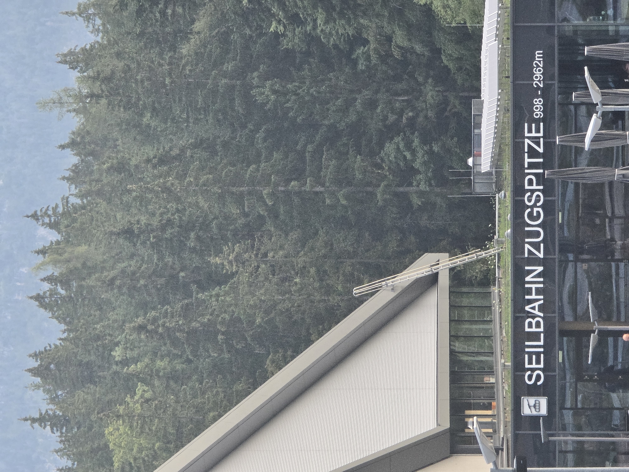 The image shows the Zugspitzbahn cable car station in Grainau, Germany. The station is a modern building with a slanted, silver-colored roof and large glass windows. Above the windows, the text "SEILBAHN ZUGSPITZE 998 - 2962m" is displayed in white lettering. The station is surrounded by a dense forest of evergreen trees, with a hazy blue sky in the background. A metal ladder leans against the side of the building. In front of the station, there are several streetlights and closed umbrellas, suggesting an outdoor seating area.