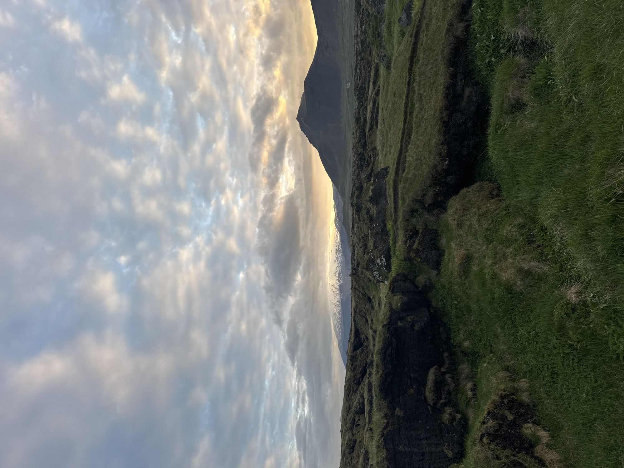 This media file captures a majestic natural landscape, likely in Búðir, Iceland, characterized by mountains, rugged terrain, and a dramatic sky. The image appears to have been taken during either sunrise or sunset, as indicated by the warm, golden light illuminating sections of the sky.
The upper portion of the frame is dominated by a dynamic sky filled with scattered clouds. These clouds range in color from soft grey to bright white and are spectacularly lit by the low sun, with hues of gold and peach reflecting off their surfaces, interspersed with visible patches of pale blue sky.
In the mid-ground, a dark, imposing mountain or large hill stands prominently on the right, its silhouette sharply defined against the radiant horizon. Further in the distance, a long range of mountains extends across the landscape, with several peaks clearly capped with snow, suggesting a high elevation or a cool climate.
The foreground consists of rugged, dark terrain, likely volcanic rock, densely covered with vibrant green grass and other hardy vegetation. A visible gully or fissure runs through this area, adding texture and depth to the immediate surroundings.
There are no people or man-made objects visible, and no activity is taking place. The scene conveys a profound sense of natural grandeur and tranquility. No text is visible in the image.
