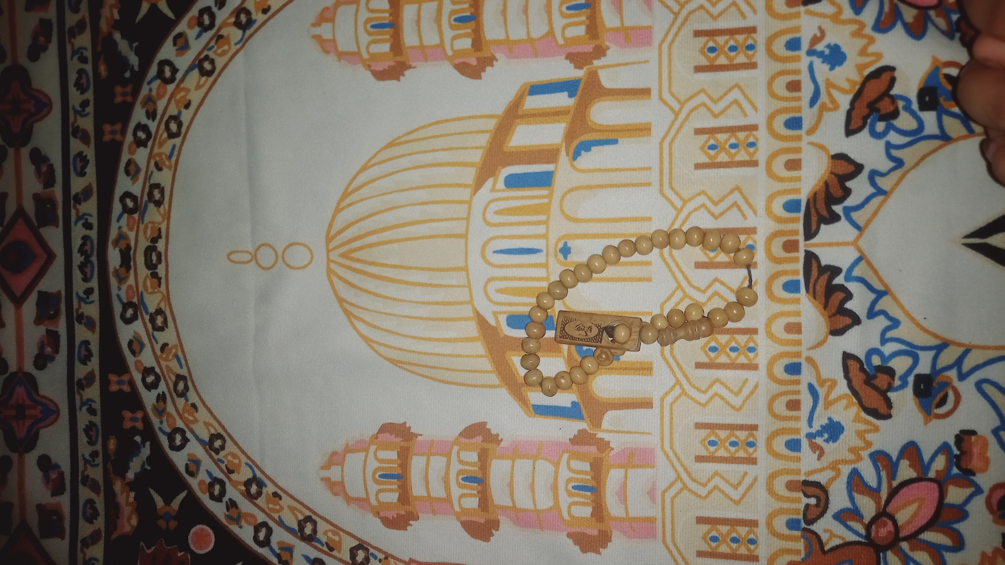 A close-up, overhead shot shows a prayer rug with an illustration of a mosque and minarets. In the foreground, a string of light-colored prayer beads lies diagonally across the rug. The prayer rug features intricate geometric patterns and floral motifs in warm tones, including shades of yellow, orange, brown, blue, and pink. The image was taken in Leuwisadeng, Indonesia.
