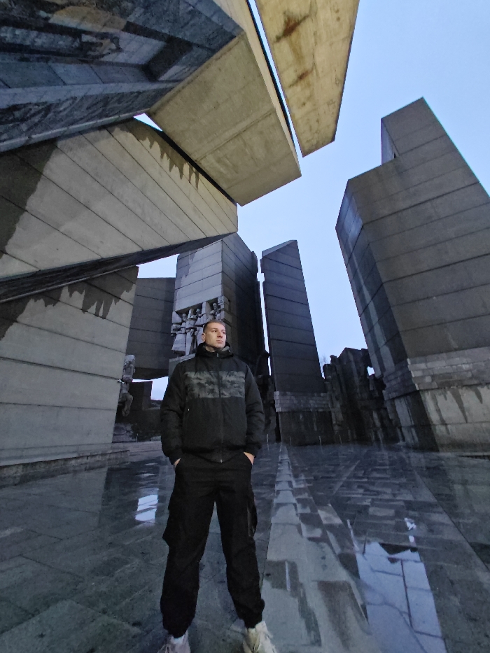A lone male stands centrally within the Monument to 1300 Years of Bulgaria in Shumen, Bulgaria. The subject is an adult, positioned facing slightly to the left, with both hands in the pockets of his black cargo pants. He wears a black hooded puffer jacket featuring a grey and black camouflage pattern across the chest and shoulders, paired with light-colored athletic shoes. His short, dark hair is visible, and his gaze is directed slightly upwards and to the left. The background is dominated by the monumental Brutalist architecture of the concrete structures, composed of large, angular grey concrete slabs and blocks, some exhibiting carved relief figures. The paved ground surface is wet and highly reflective, showing clear specular reflections of the surrounding structures and the pale blue sky above. The image is captured from a low-angle perspective, emphasizing the scale of the monument.