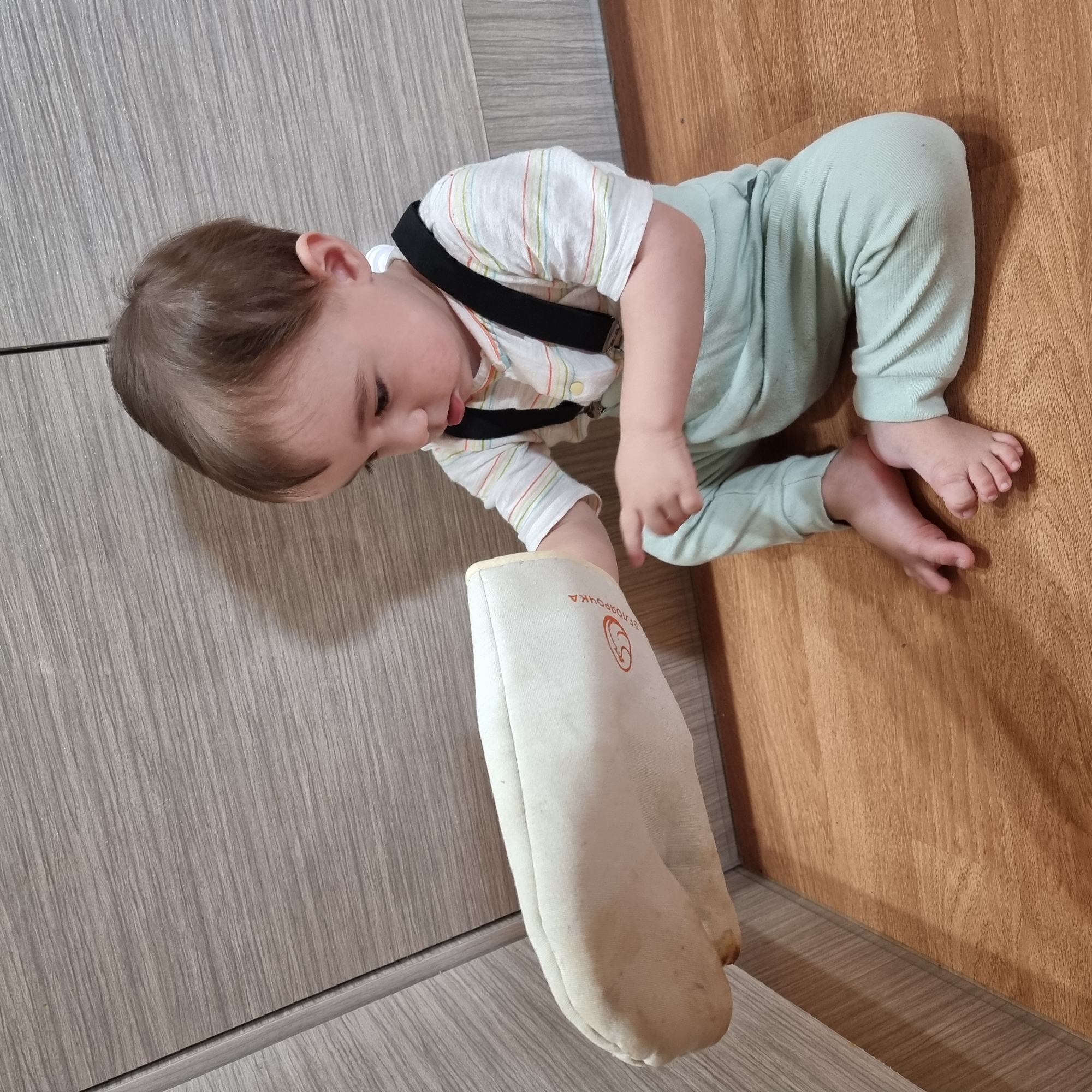 A young child, appearing to be between one and two years old, is seated on a wooden floor in what seems to be a kitchen or utility area. The child is wearing a light blue long-sleeved outfit with suspenders and a horizontally striped shirt. In their left hand, the child is holding a light-colored, padded object, possibly a pot holder or oven mitt, which has an orange circular logo with text. The child is looking down at the object, with their tongue slightly out. The background consists of a wall with a wood-grain pattern, likely cabinets or paneling, and the floor is made of polished wood planks. The scene appears to be an indoor domestic setting.