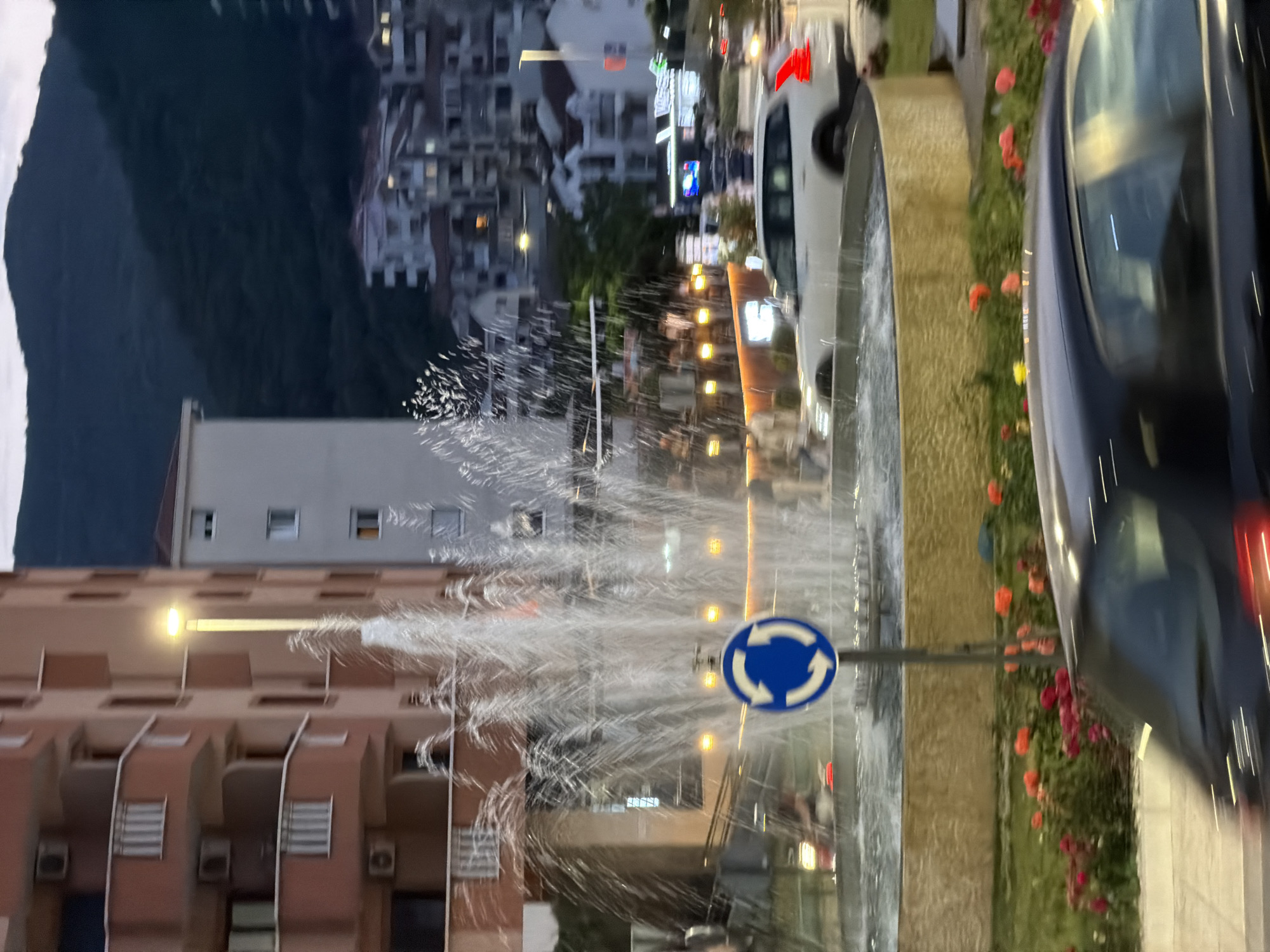 The image depicts an urban scene in Budva, Montenegro. In the foreground is a roundabout featuring a fountain. The fountain's water jets are active. A blue and white roundabout traffic sign is clearly visible. A dark-colored car is parked nearby, partially obscuring the view. The background shows several buildings, including residential blocks with visible windows and balconies. In the distance, there's a mountain under an overcast sky. Street lighting illuminates the scene. There are also other cars on the road.