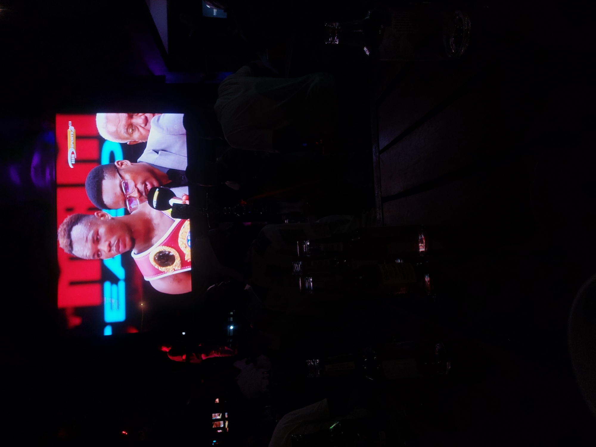 The image shows a television screen displaying a sports broadcast, possibly boxing-related given the presence of a boxer with a championship belt. The broadcast is likely from SuperSport, as indicated by the visible logo. The screen is set up in what appears to be a bar or pub setting, as suggested by the tables, glasses, and shadowy figures of people in the foreground. Specific details about the location within Dar es-Salaam, Tanzania, are not evident from the given visual information.