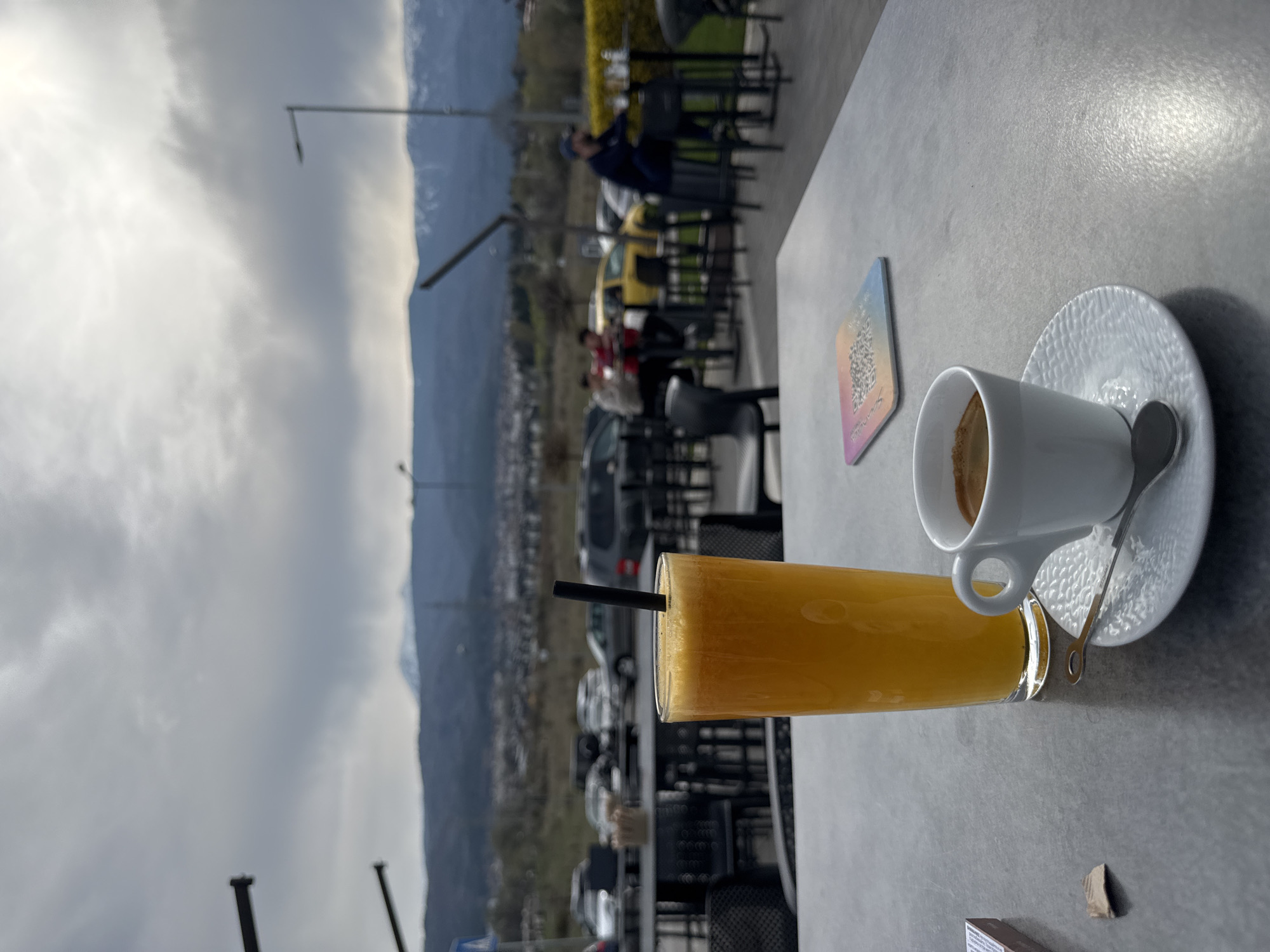 On a grey, textured table, a glass of orange juice with a black straw is positioned alongside a white coffee cup on a textured saucer with a silver spoon. A card with colorful graphics rests near the beverages. The table appears to be part of an outdoor seating area, with black chairs and tables stretching into the background. Beyond the seating area, a city extends towards the base of a mountain range, partially obscured by a cloudy sky. The scene may be near or in Sofia, Bulgaria, but I can not be sure. Several people are seated in the background. Cars are also visible.