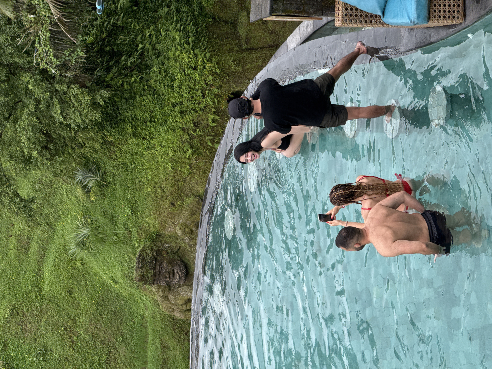 The image shows an outdoor scene in Tegallalang, Indonesia, featuring a curvilinear pool with multiple people. A couple, the man in a black t-shirt and cap and the woman in a black swimsuit, are standing at the edge of the pool, interacting with the water. Further into the pool, another couple is seen; the man is shirtless, wearing dark swim trunks, and the woman is in a red swimsuit. The man is holding a smartphone, possibly taking a photo. The pool's design includes strategically placed stone platforms within the water. Adjacent to the pool's edge, there is dense, lush greenery. A section of a woven chair with blue cushions is visible in the upper-right corner.