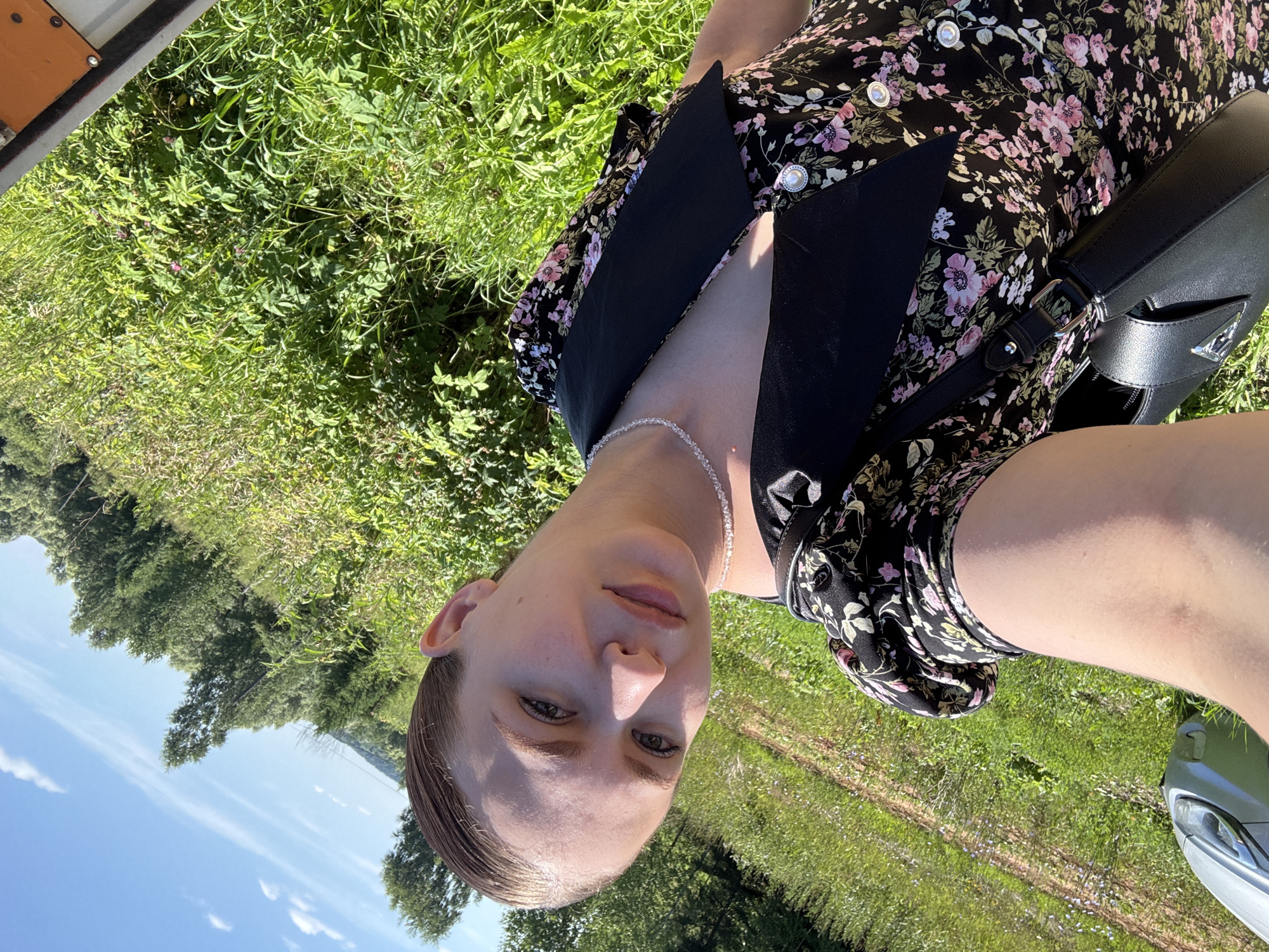 The image shows a young person in a floral dress sitting outdoors on a sunny day. The scene appears to be a field or meadow, with lush green grass and trees in the background. The person is looking towards the camera, with a slight smile. They are wearing a black dress with a floral pattern of pink and white flowers. A black purse is slung over their shoulder. The sky is blue with a few scattered clouds, suggesting pleasant weather. The image is taken from a slightly elevated angle, looking down at the subject. The lighting suggests it is daytime, likely late morning or early afternoon due to the bright sunlight. The location is identified as Nizhnyaya Gar', Russia. There are no visible text elements in the image.