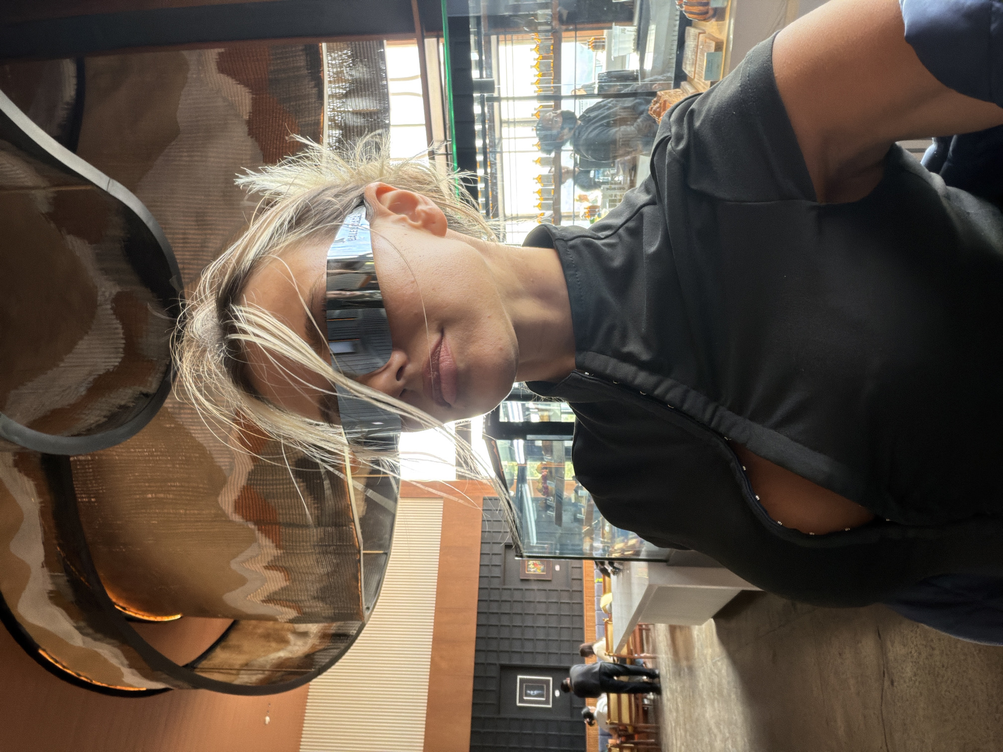 A close-up, selfie-style photograph shows a woman with blonde hair and futuristic silver-tinted sunglasses. She is wearing a black top with a high neckline and an open front, revealing her chest. Her hair is messy and windswept. The background suggests an indoor setting, possibly a cafe or restaurant, with a large, decorative light fixture overhead. In the middle ground, there are tables and chairs with people seated, and a display case with food items. A man in a black jacket is visible in the background, as well as shelves with bottles. The overall lighting is warm.