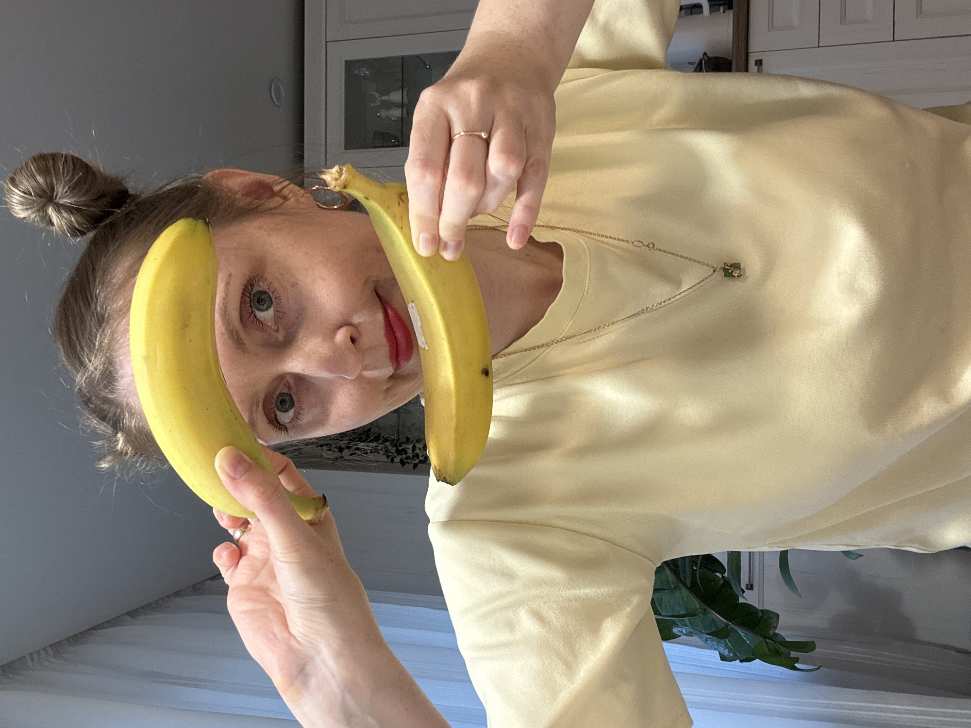 A woman with blonde hair in a bun is holding two bananas, one in each hand, near her face. She is wearing a light yellow shirt and a gold necklace with a pendant. She has red lipstick on and a ring on her finger. The background includes a white cabinet and a plant. The photo is taken indoors. The location and city of Chaikovsky, Russia is not identifiable in the provided image.