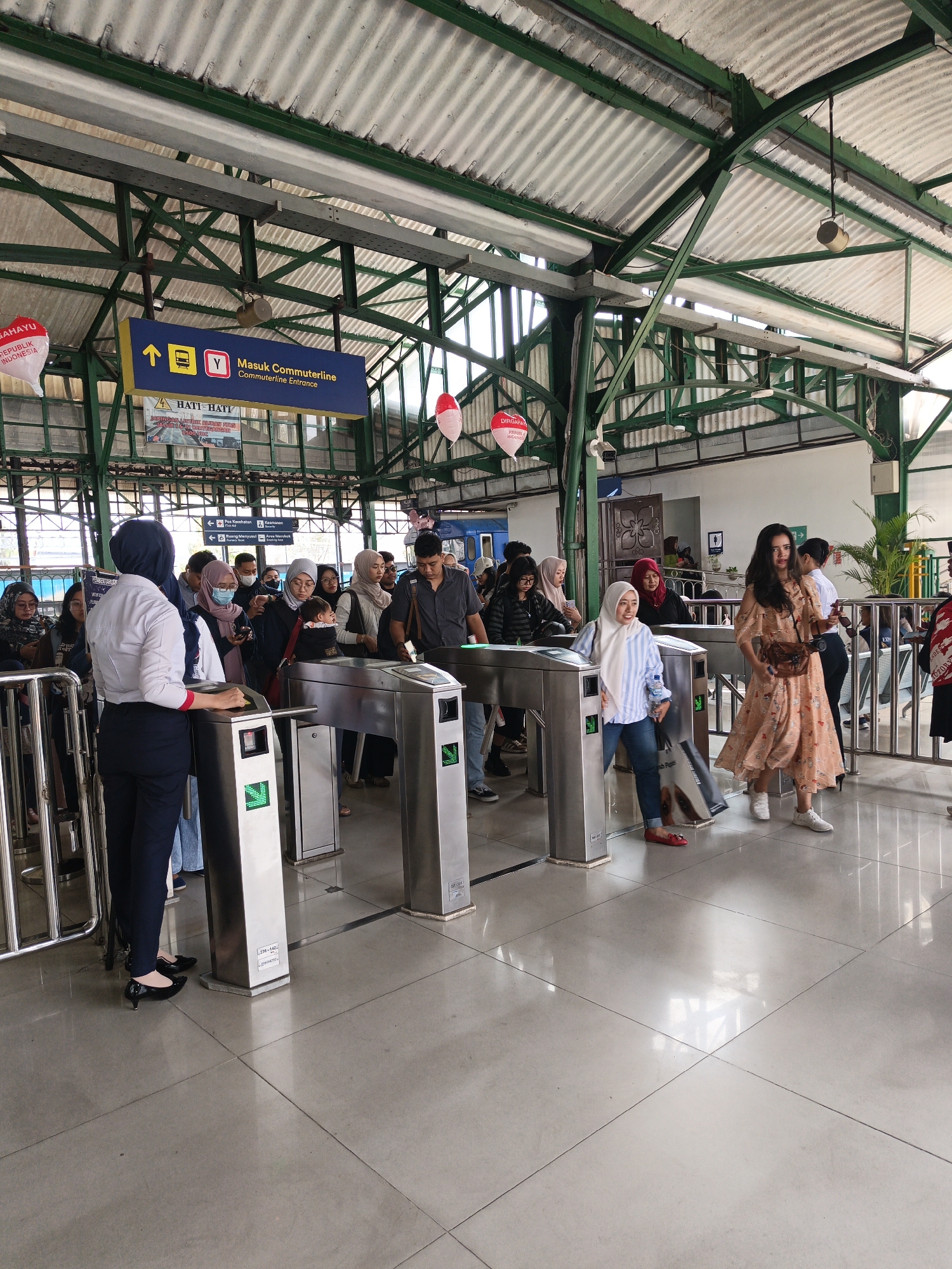 This is a wide shot of a commuter line entrance at a train station in Surakarta, Indonesia. The scene is outdoors, under a green metal roof supported by green metal beams. There are several silver ticket gates in the foreground, with people lining up to pass through them. Some individuals are wearing masks. A woman in a white shirt and dark pants, presumably a station attendant, stands near one of the gates, facing the line of people. There are signs above the gates, one of which reads "Masuk Commuterline" with an arrow and train icon. Red and white balloons are hanging from the ceiling. In the background, a blue train can be seen. The floor is tiled with light grey tiles. The overall atmosphere appears to be busy, with many people waiting to enter the commuter line.