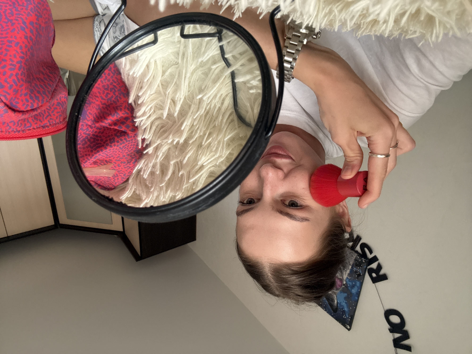 A young woman with her hair pulled back in a bun is applying makeup in front of a mirror. She is holding a red makeup brush to her cheek. She is wearing a white t-shirt and a silver watch. The mirror is reflecting a fuzzy white rug and a red and purple patterned object, possibly a makeup bag. In the background, there are shelves and a wall with some decorations. The scene appears to be indoors, possibly in a bedroom or bathroom. There is no indication of the location being in the city of Perm, Russia.