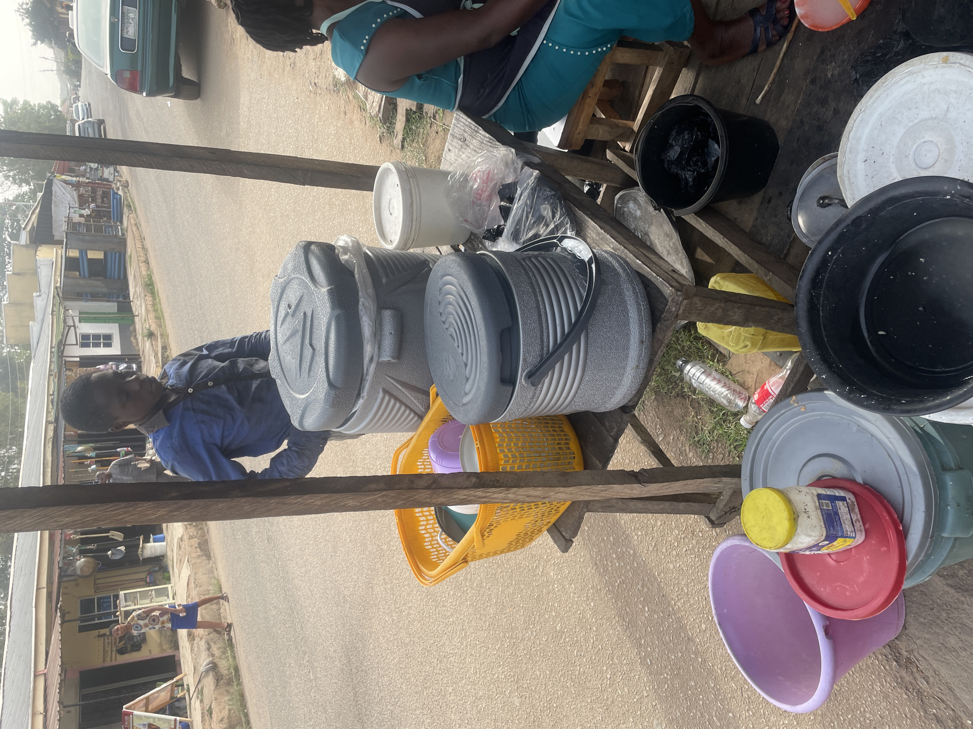 The image shows a roadside vendor in Jos, Nigeria, selling cold drinks. Two large insulated containers, grey in color, dominate the scene, presumably filled with beverages. A yellow basket is positioned below them, holding various plastic containers, including a purple bucket and some covered containers. The vendor, wearing a blue shirt, stands to the left of the containers. A woman, wearing a teal and navy top, sits on a stool to the right. Assorted plastic buckets and containers are arranged on the vendor's table. In the background, a street scene is visible, showing buildings and vehicles along a paved road.