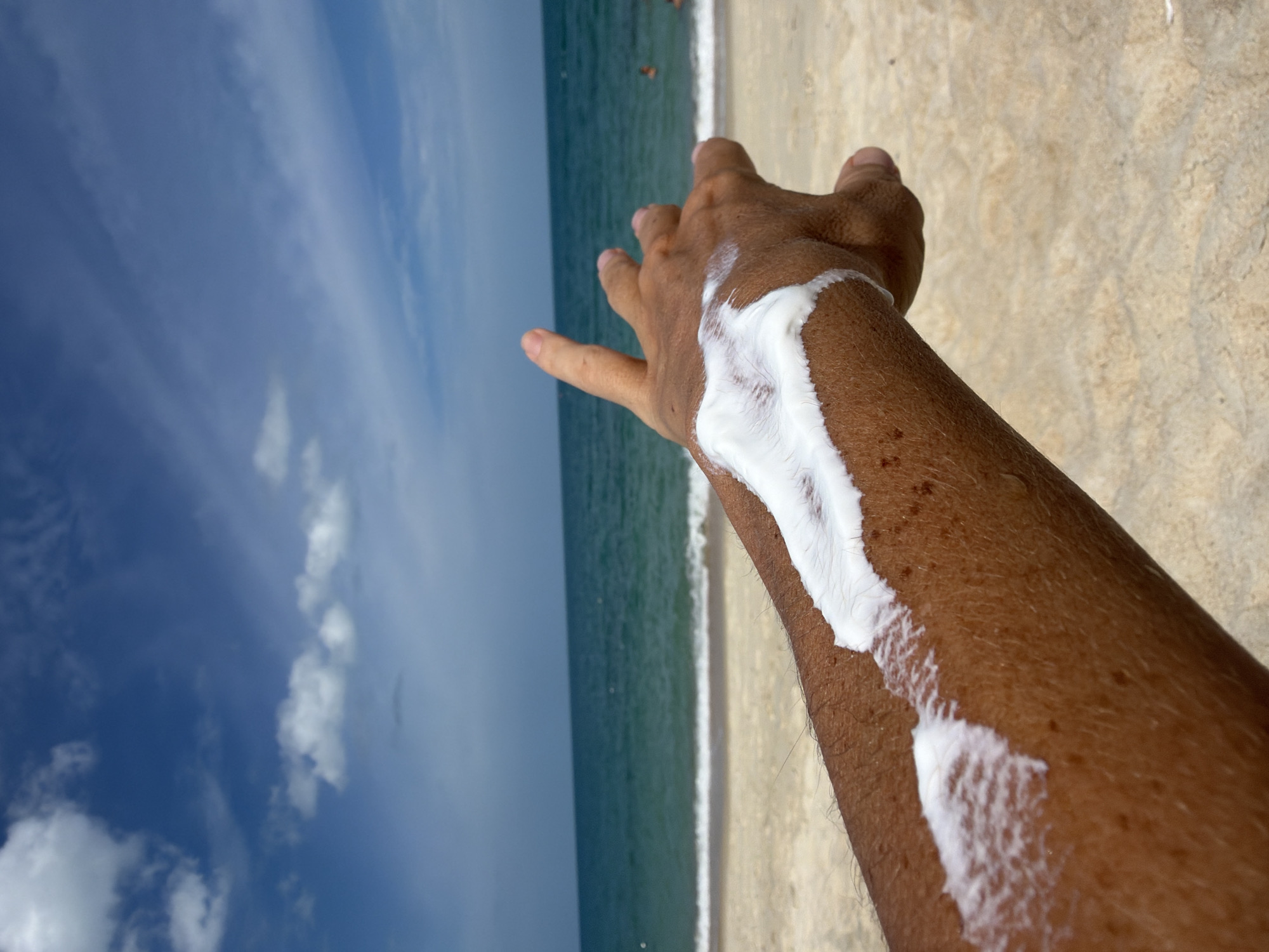 A person's forearm, with sunscreen applied in a thick white stripe, is held up against a beach scene. The background consists of a sandy beach, the turquoise ocean, and a blue sky with scattered white clouds. The scene is a close-up shot focusing on the arm and the sunscreen, with the beach and ocean serving as a backdrop.