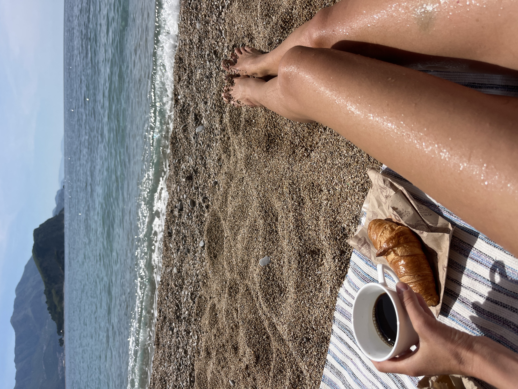 A woman sits on a pebble beach in Budva, Montenegro, beside the Adriatic Sea. She has a cup of black coffee in her hand and a croissant in a paper bag on a striped towel next to her legs. The turquoise water is visible to the left, and a large forested mountain is visible in the background. The woman's legs are suntanned and appear wet.