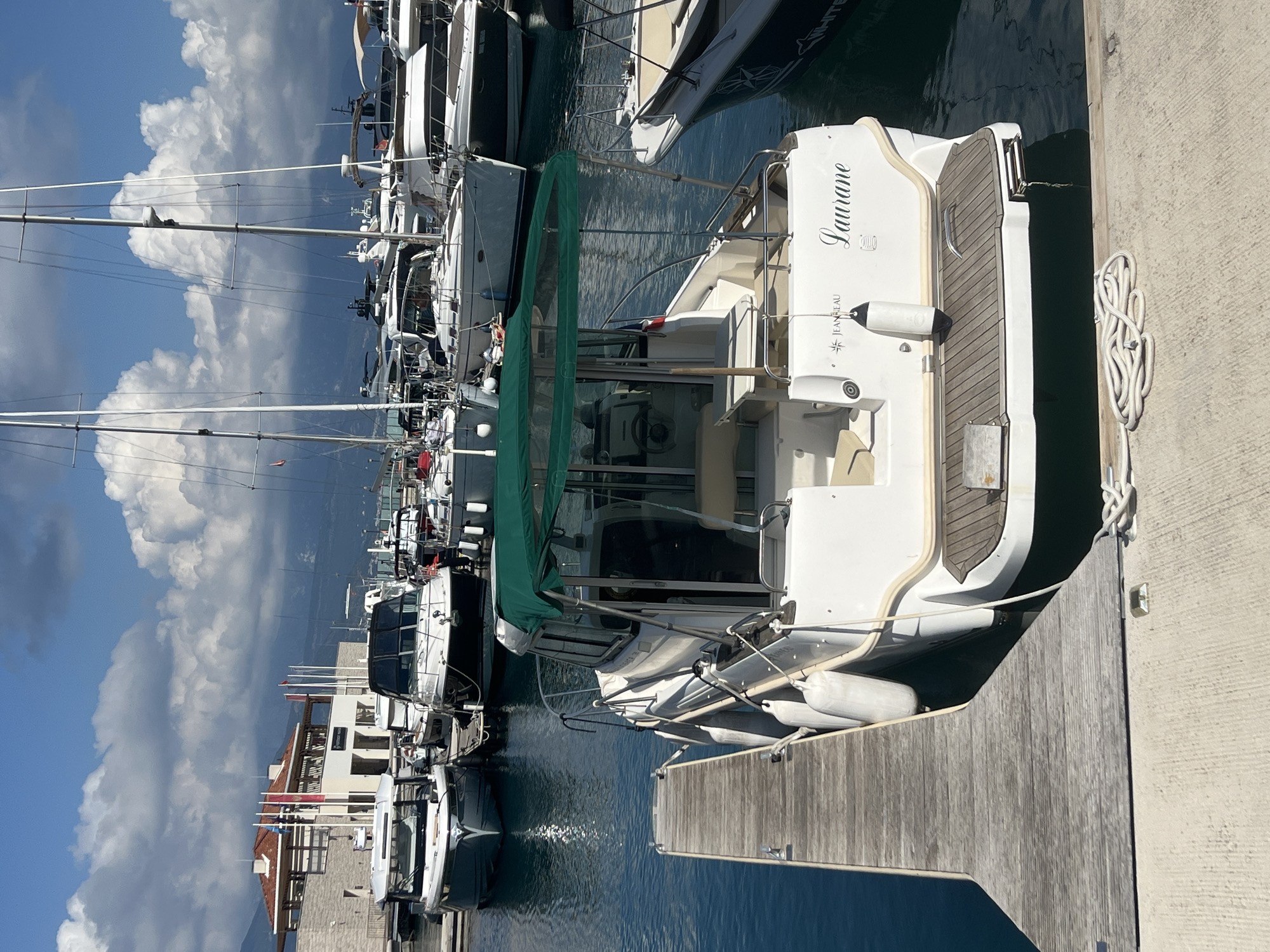 This image captures a serene marina scene in Đenovići, Montenegro, on a bright, sunny day. The sky is a clear blue with large, fluffy white cumulus clouds.
In the foreground, a white motor yacht named "Laurane" is prominently docked. The name "Laurane" is written in an elegant script font on its stern, with "JEANNEAU" and a logo (likely "SANREMO") visible below it, indicating the boat's manufacturer and model. This yacht features a green canvas cover over part of its cockpit area and has several white fenders hanging along its side. Its stern has a wooden deck area.
The yacht is moored to a weathered wooden dock, on which a white rope is neatly coiled. The water in the marina is calm, reflecting the boats and sky, with slight ripples visible from the gentle movement of the water.
In the background, numerous other boats, including both sailboats with tall masts and other motor yachts, are docked along the marina. Beyond the boats, a cluster of buildings with terracotta roofs and light-colored facades can be seen, hinting at the coastal architecture of the area.
The scene conveys a peaceful and leisurely atmosphere, typical of a marina environment under pleasant weather conditions.