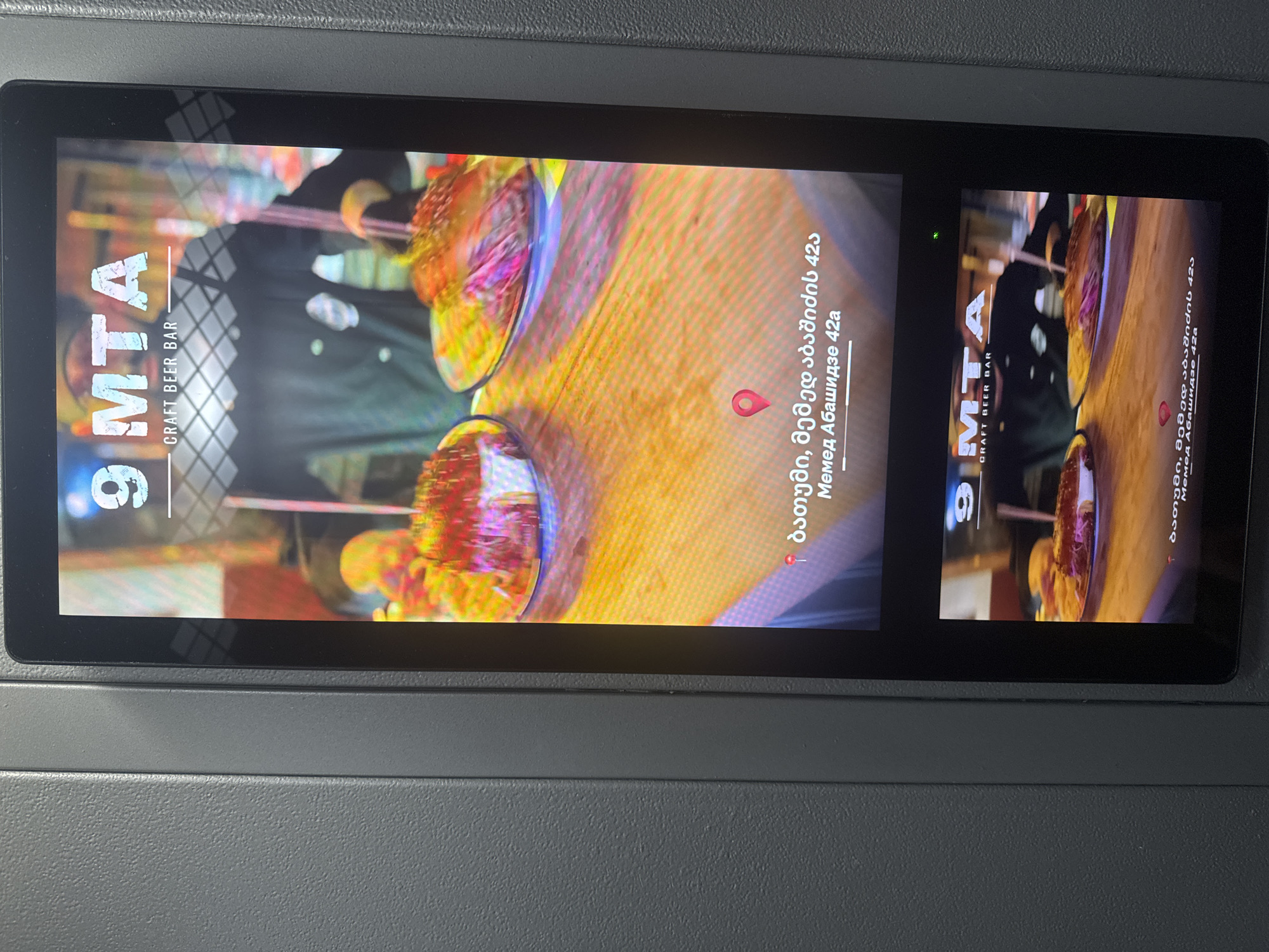 A vertical digital display screen is depicted, embedded within a textured, dark grey panel. The screen exhibits an advertisement for "9 MTA CRAFT BEER BAR." The prominent upper portion of the display features an image of two gourmet burgers or similar dishes on plates, presented on a wooden surface. A blurred background shows a person in a dark uniform or shirt. Below this imagery, text in Georgian is displayed, indicating "ბათუმი, მემედ აბაშიძე 42ა" (Batumi, Memed Abashidze 42a), accompanied by a red location pin icon. A smaller, repeating version of this advertisement is visible at the bottom of the screen. A small green indicator light is present on the lower right bezel of the display. The location identified is Batumi, Memed Abashidze 42a.