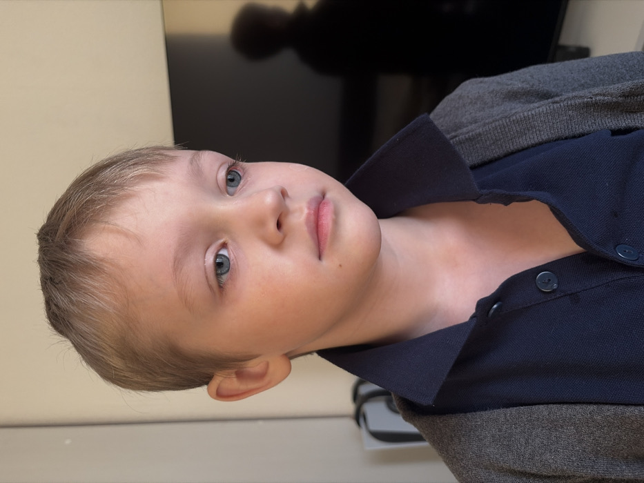 A young Caucasian male child, estimated to be between five and eight years old, is depicted in a close-up, chest-up indoor portrait taken in Saint Petersburg, Russia. He has short, light brown hair with bangs, blue eyes, and fair skin, with a faint redness observed under both eyes. A small mole is visible on his right cheek, below the nostril. His gaze is directed slightly downwards and to his right, with a neutral to pensive expression. He is wearing a dark blue polo shirt with an open collar and two visible dark blue buttons, layered beneath a grey knitted cardigan. The background on the left consists of a plain light-colored wall, with a partial view of a grey and white object, possibly a bag or console, behind his left shoulder. On the right, a large dark rectangular surface, consistent with a television screen, reflects a vague dark silhouette of a person. The lighting is even and consistent with an interior setting.